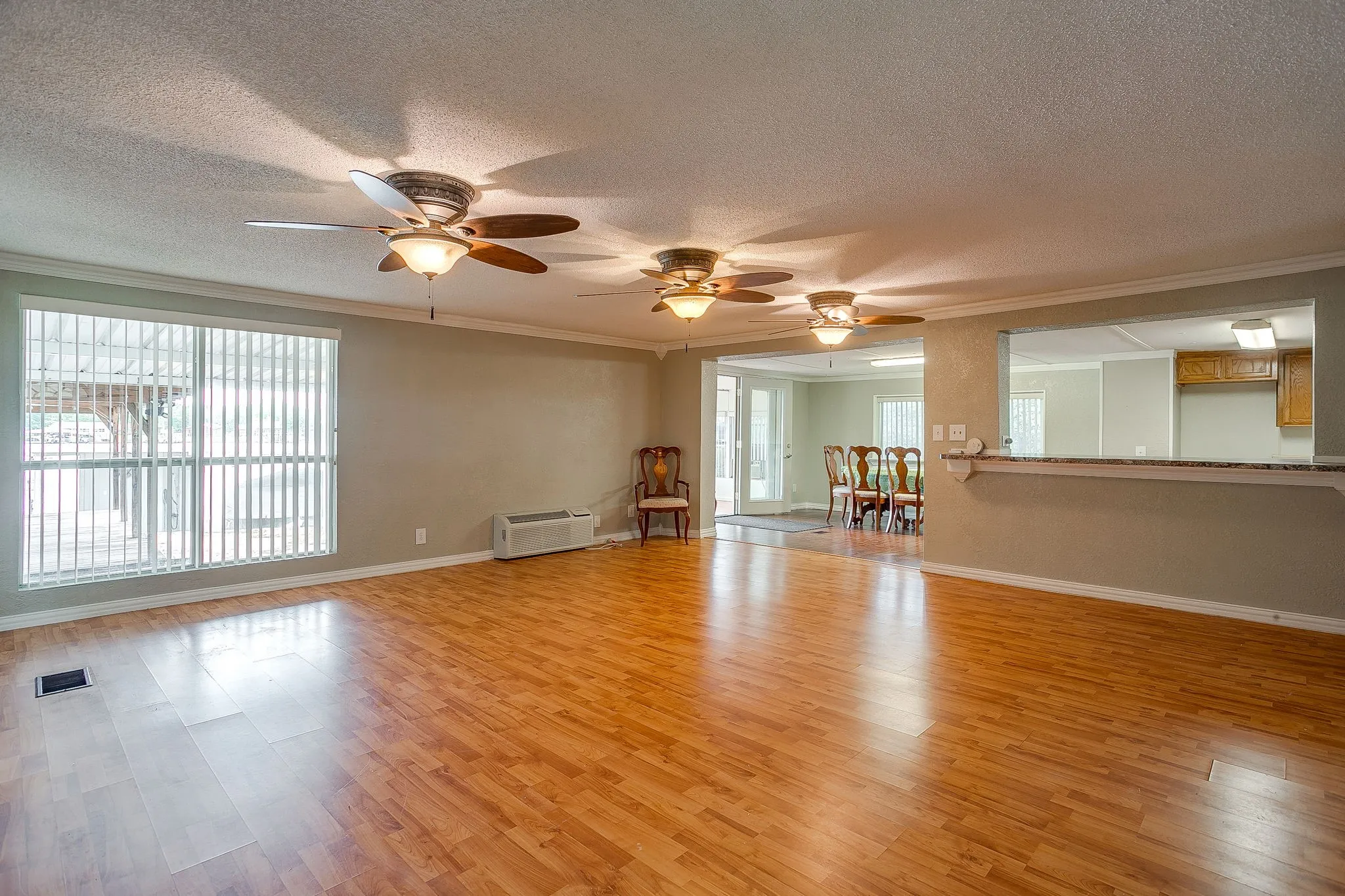 Spacious living area featuring wood-finish flooring, multiple ceiling fans with integrated lighting, a large window with vertical blinds, and a pass-through counter opening to an additional room
