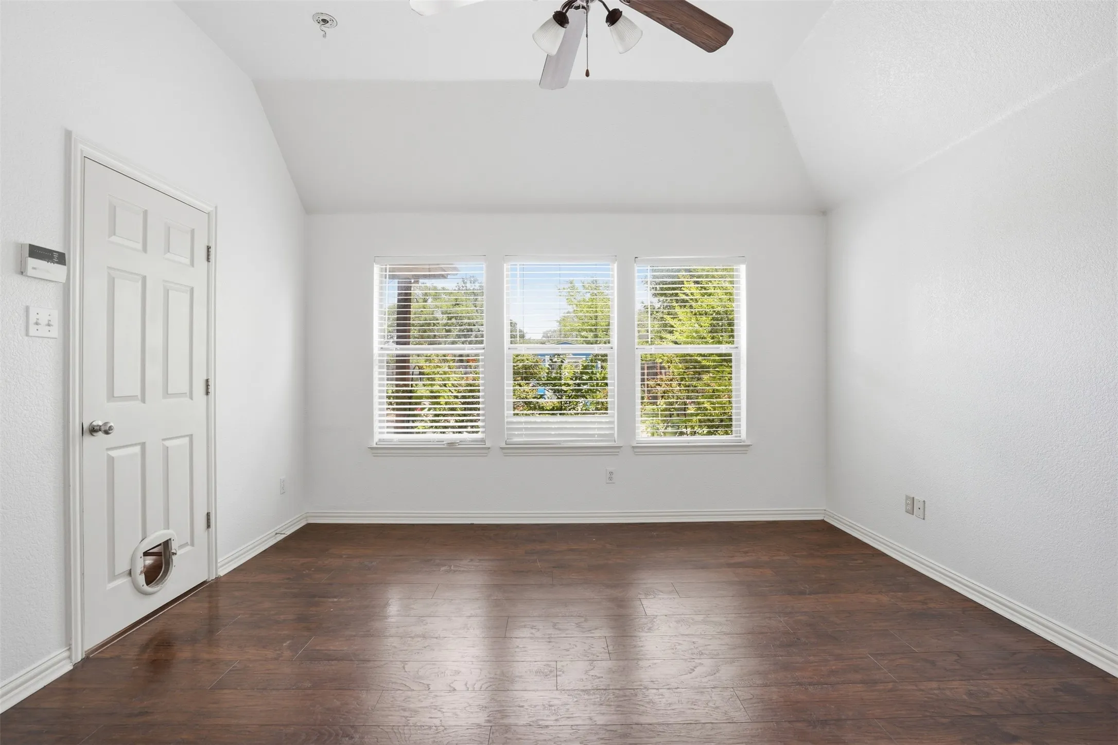 Spacious room featuring vaulted ceilings, rich wood-finish flooring, three large windows with horizontal blinds, and a ceiling fan with integrated lighting