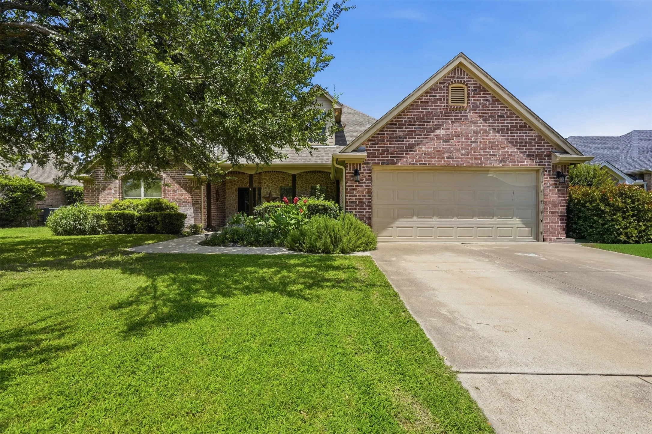 Brick exterior featuring a two-car garage, mature landscaping, and a concrete driveway