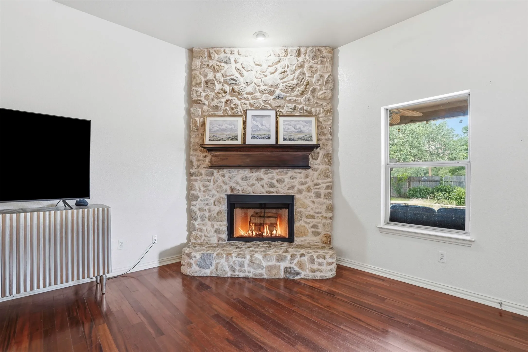 Stone-clad fireplace with a dark wood mantel, recessed lighting, wood-finish flooring, a white window frame, and white wall finishes
