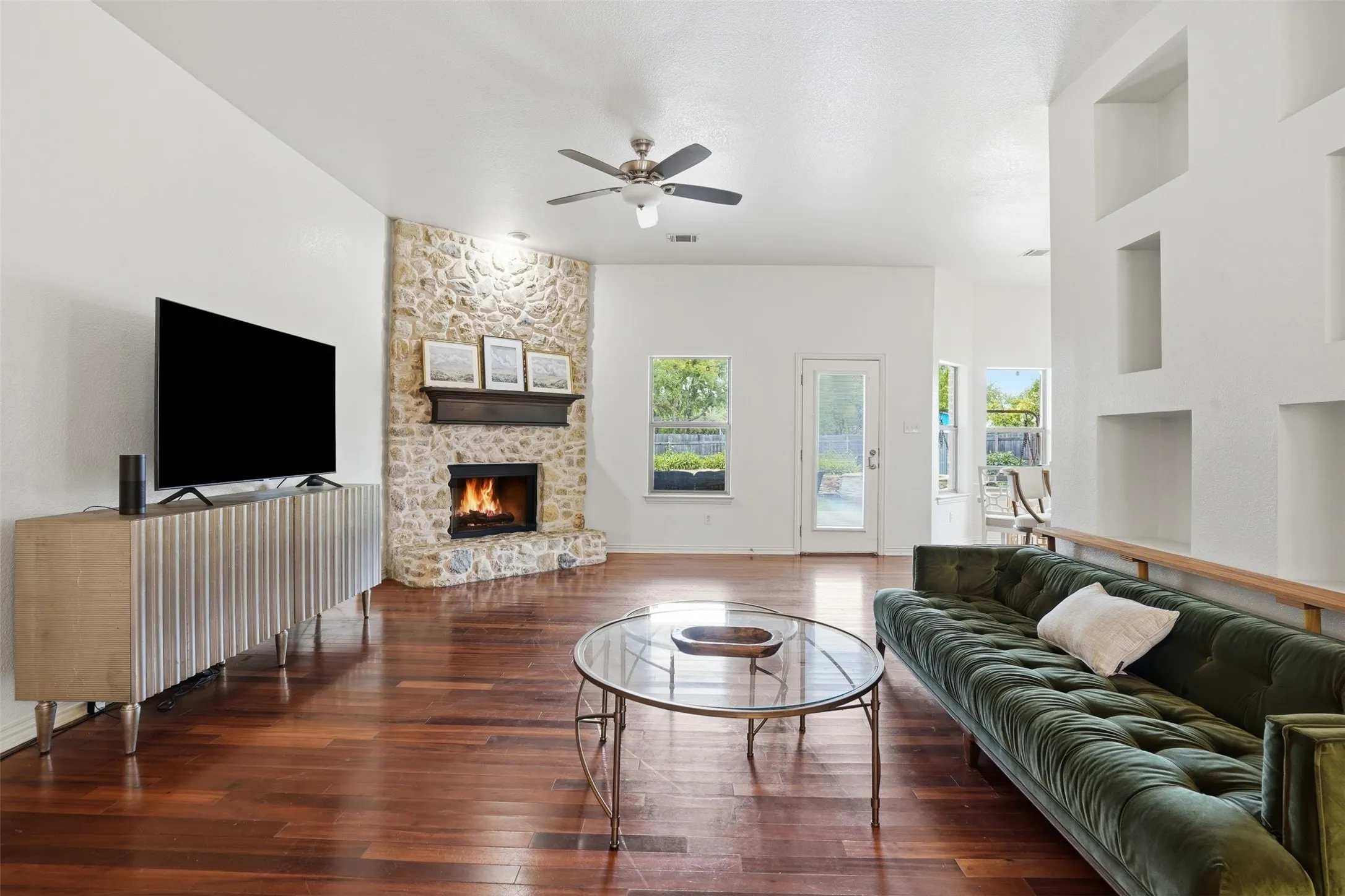 Spacious living area featuring wood-finish flooring, a corner stone fireplace with a wood mantle, a ceiling fan, multiple windows for natural light, and built-in wall niches