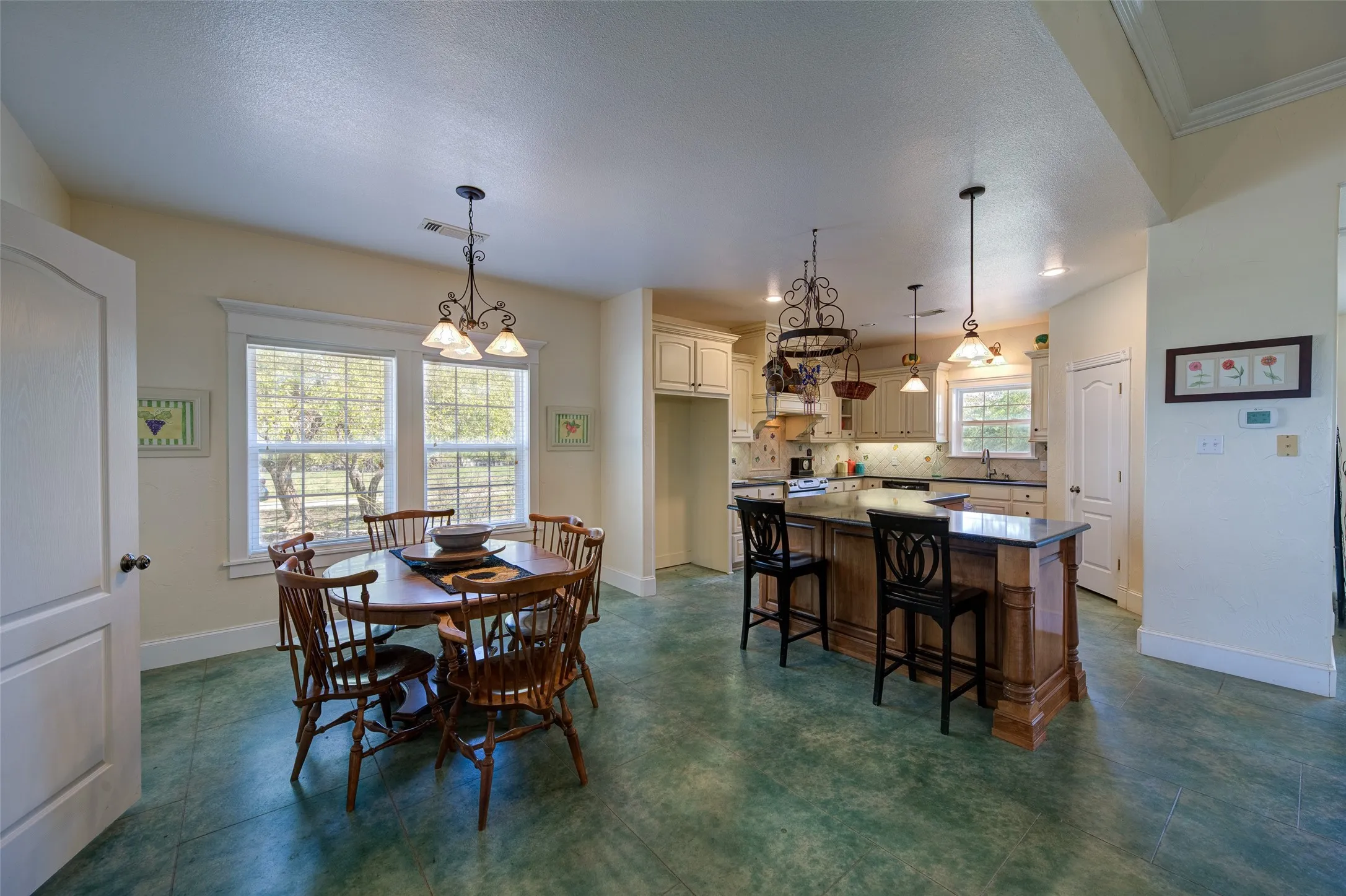 Kitchen and dining area featuring green-hued concrete flooring, white cabinetry, a large kitchen island with counter seating, and two large windows with horizontal blinds