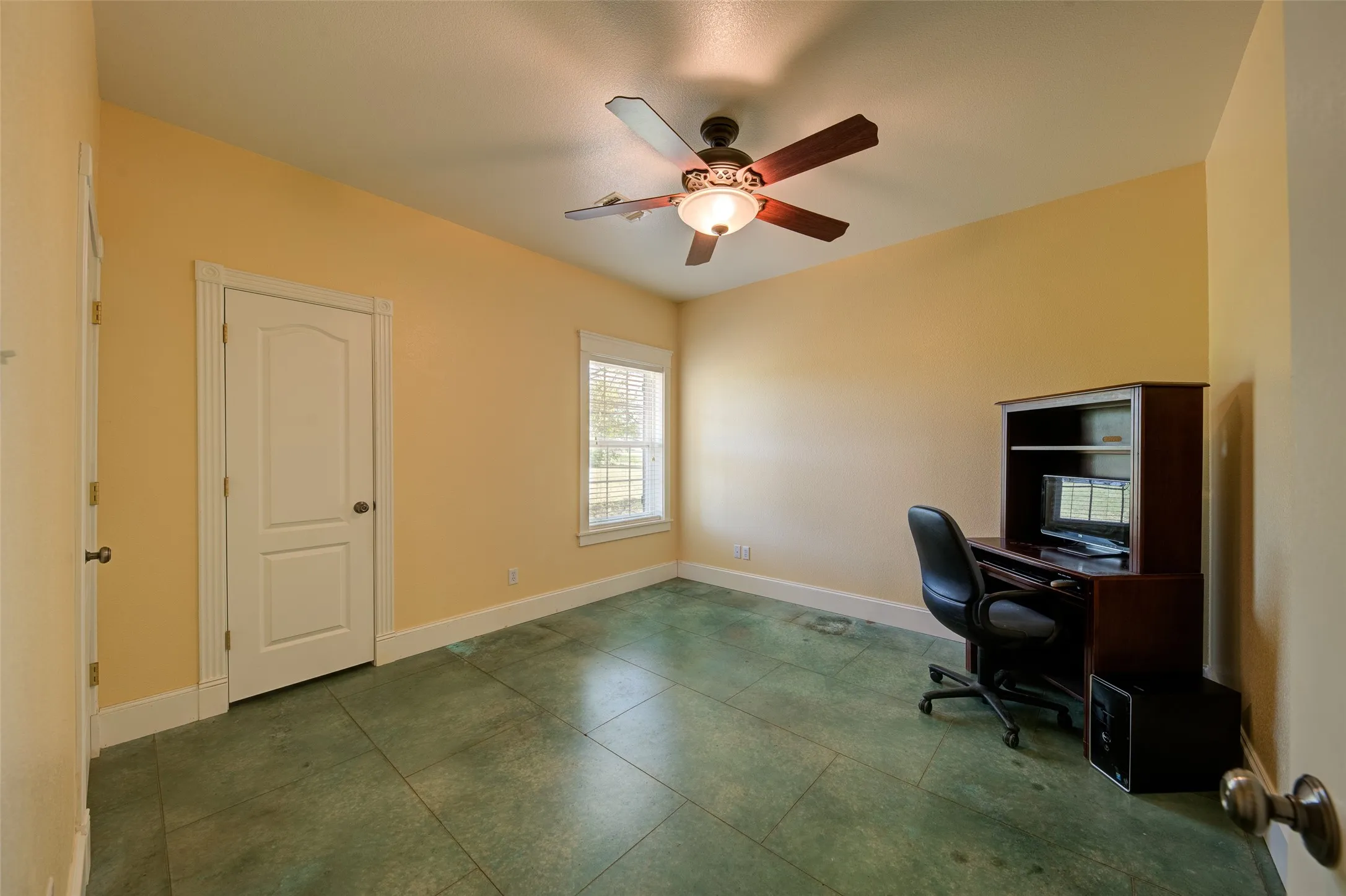 Spacious room featuring green-hued concrete flooring, a large window, a ceiling fan with integrated lighting, and white baseboards
