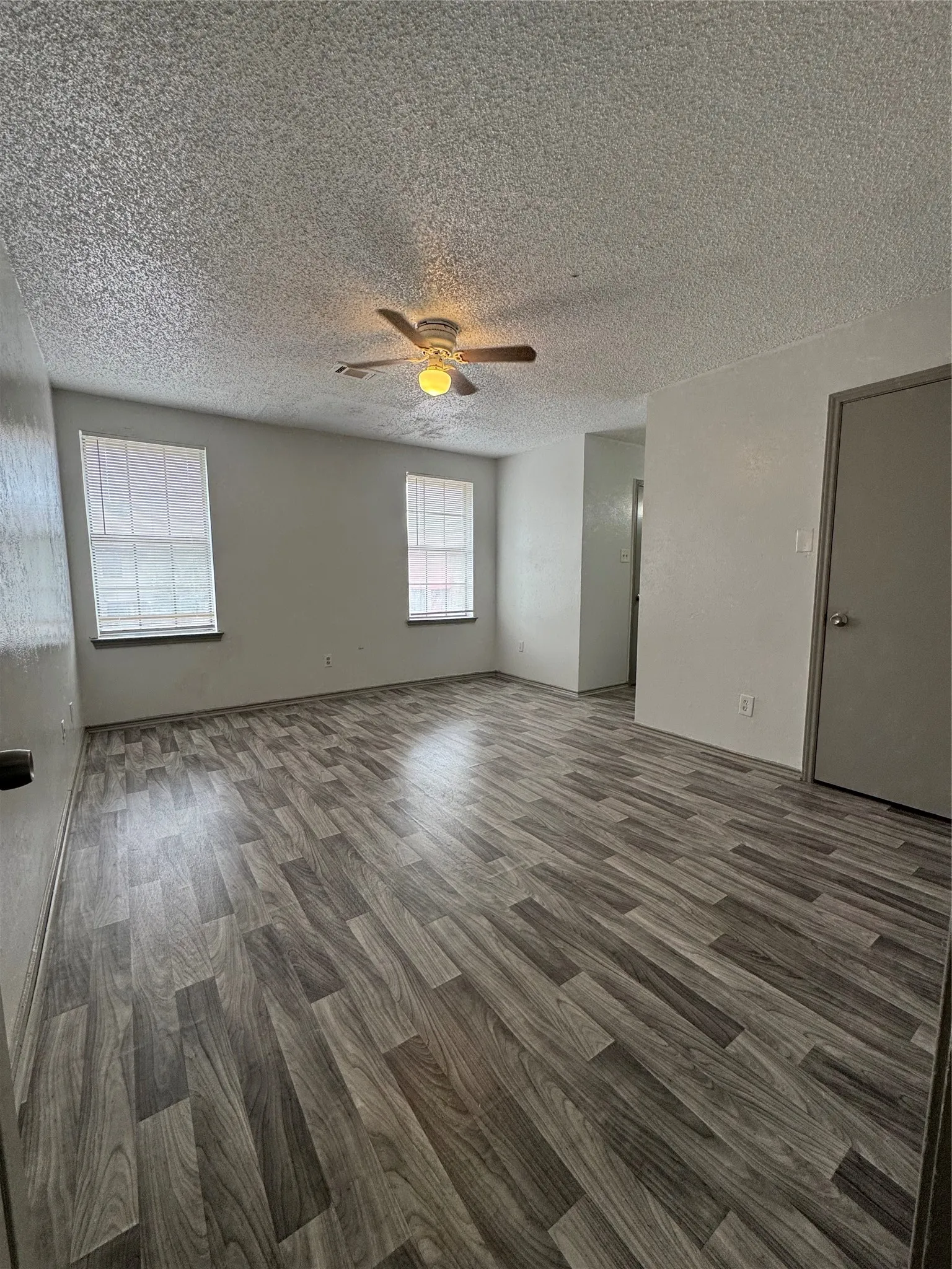Spacious room featuring wood-finish flooring, two large windows with blinds, a ceiling fan with integrated lighting, and neutral wall paint