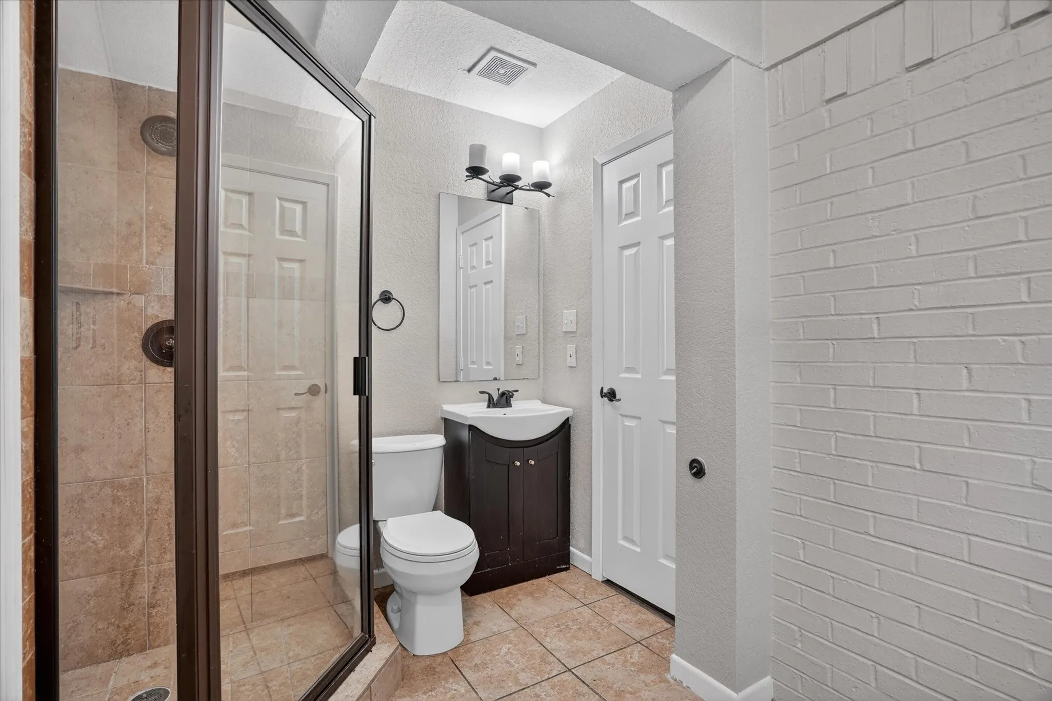 Glass-enclosed shower with tiled walls and bronze fixtures, complemented by a floor-mounted toilet, dark wood-finish vanity with a white basin, and a framed mirror under a three-light fixture