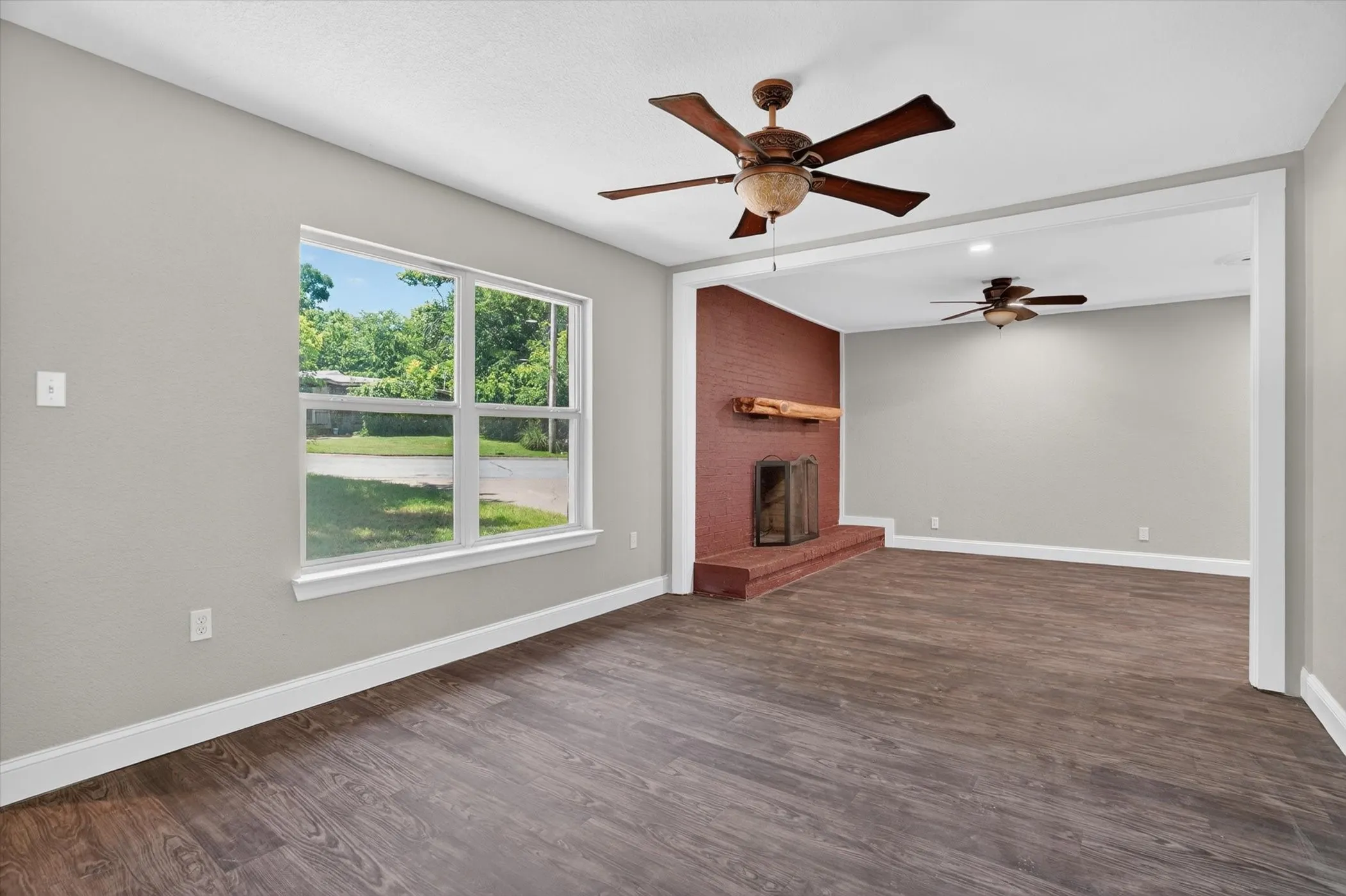 Spacious living area featuring wood-finish flooring, a red brick fireplace with a wood mantle, and a large window with white trim