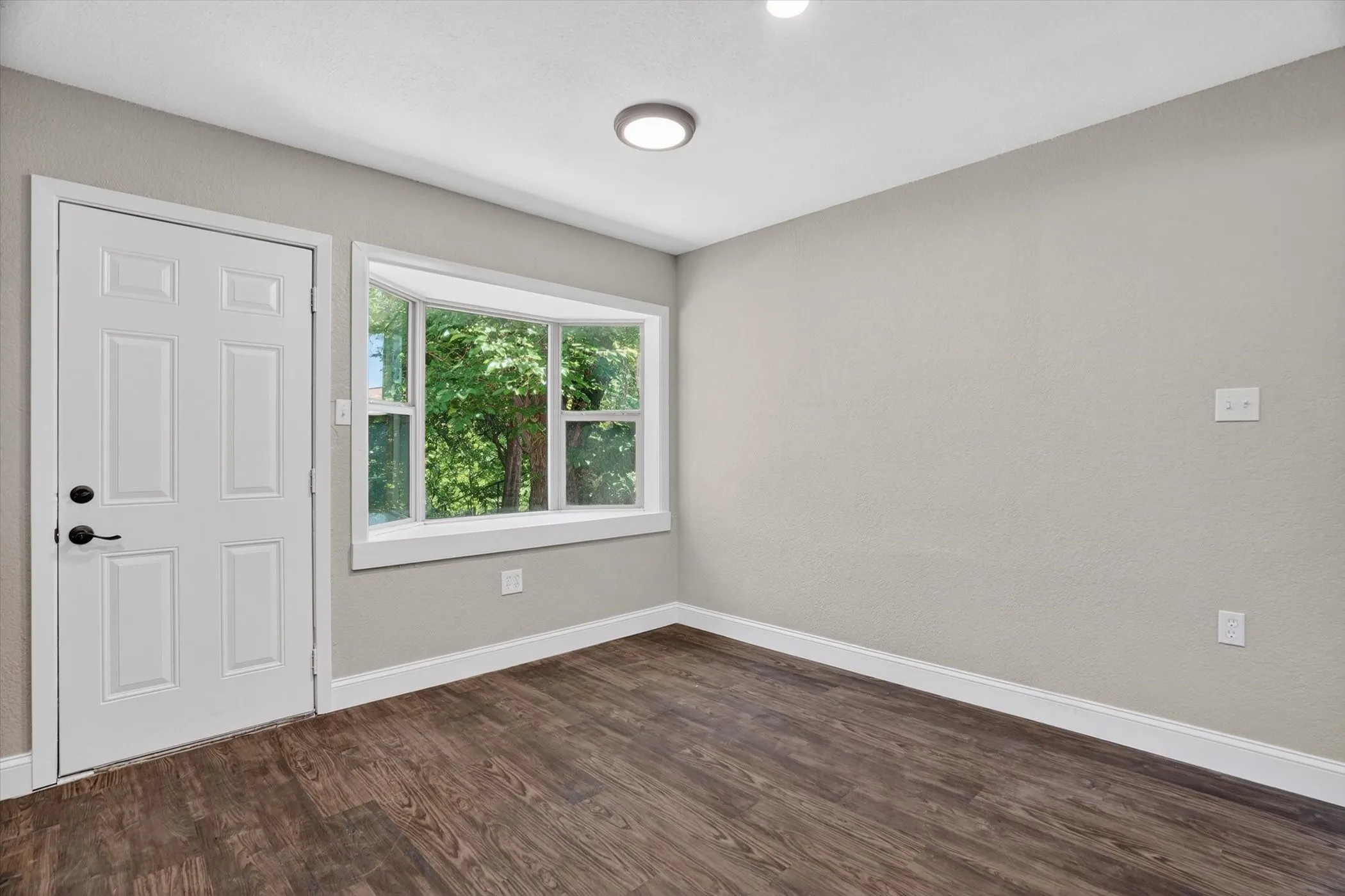 Room featuring wood-finish flooring, a multi-pane bay window, and a white six-panel door with black hardware