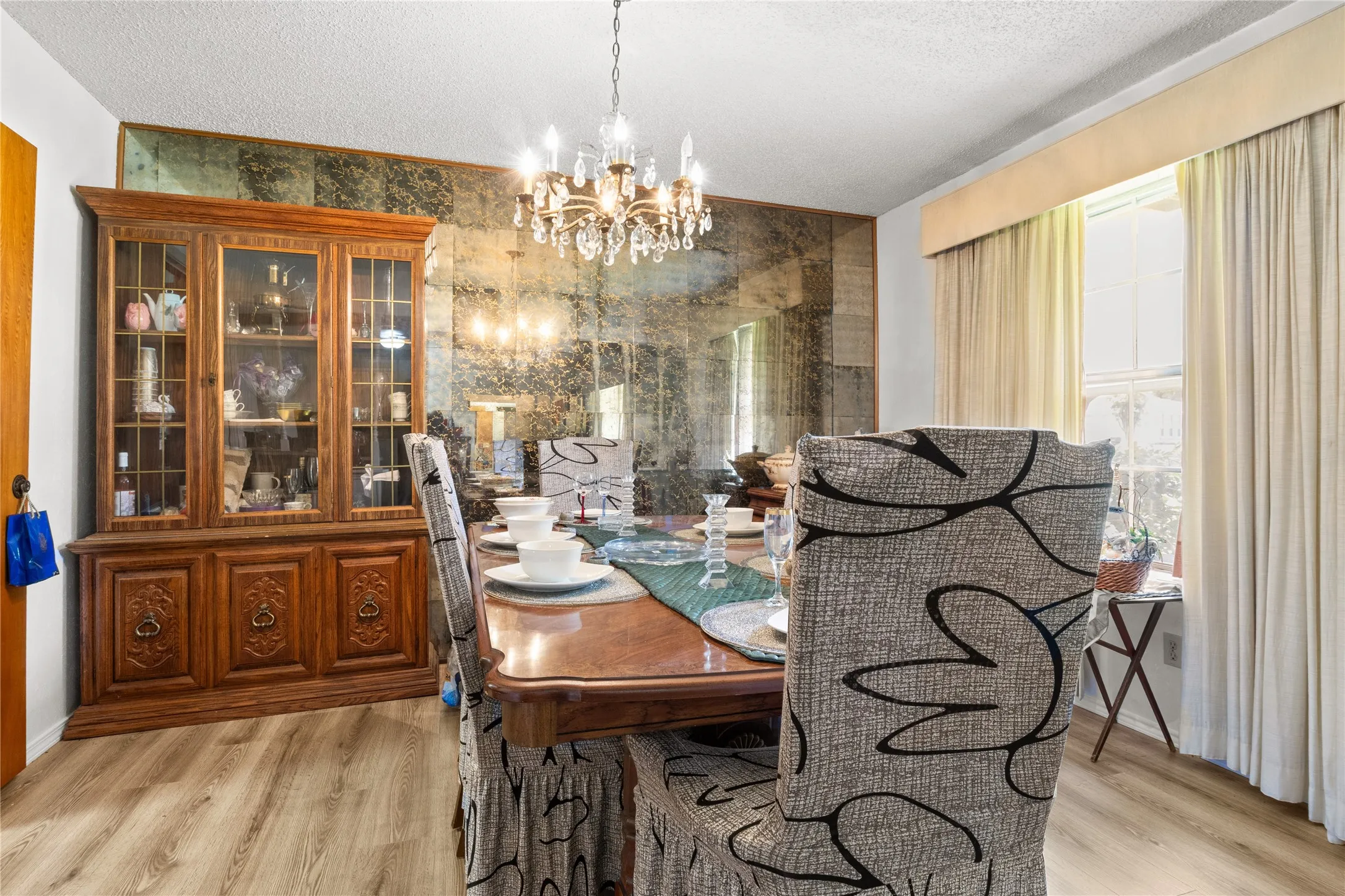 Dining space featuring an antique mirrored accent wall with integrated cove lighting, wood-finish flooring, a multi-pane window with drapery, and a chandelier with multiple glass globes
