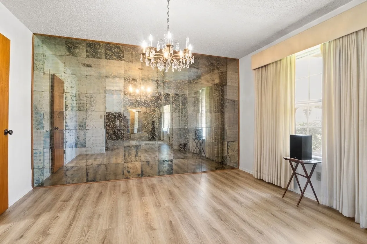 Virtually (Empty) -Dining space featuring an antique mirrored accent wall with integrated cove lighting, wood-finish flooring, a multi-pane window with drapery, and a chandelier with multiple glass globes