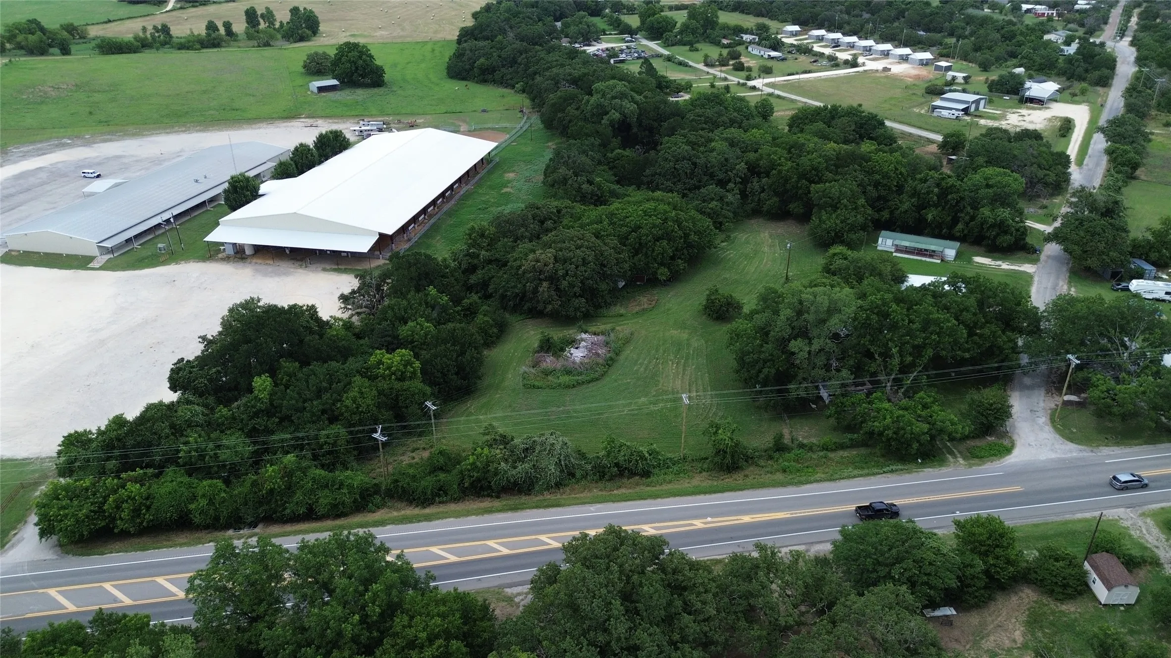 Expansive property featuring a large white roofed structure, a second gray roofed structure, a paved road frontage, and abundant mature trees