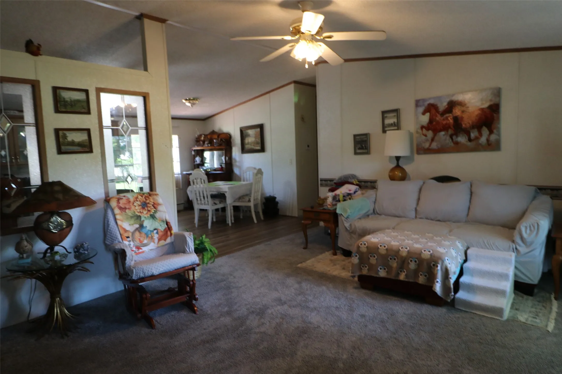 Carpeted living area featuring a ceiling fan with integrated lighting, wood-finish wall trim, and a partial decorative wall with leaded glass panels