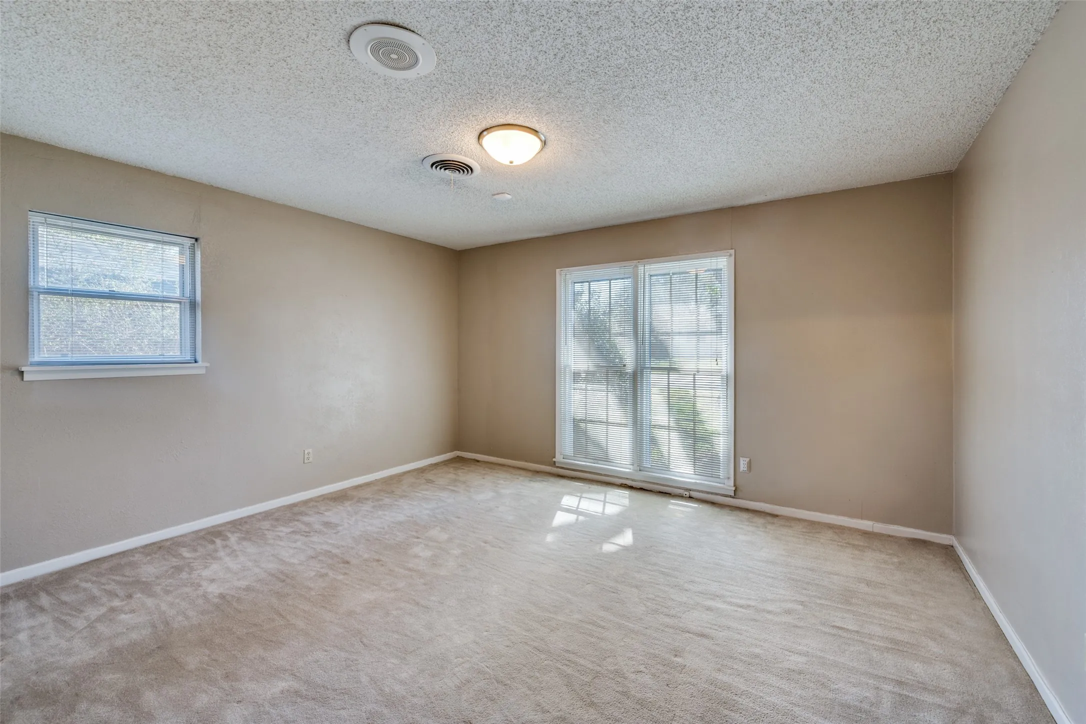 Spacious room featuring neutral carpeting, two windows, a ceiling-mounted light fixture, and a ceiling-mounted speaker