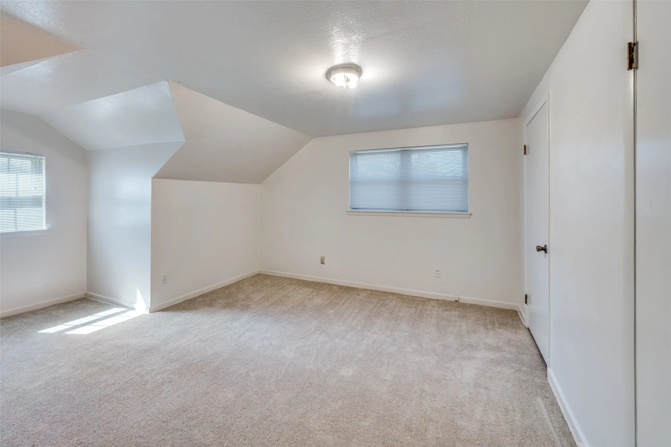 Spacious room featuring vaulted ceilings, a window with blinds, light beige carpeting, a flush-mount ceiling light, and a white interior door