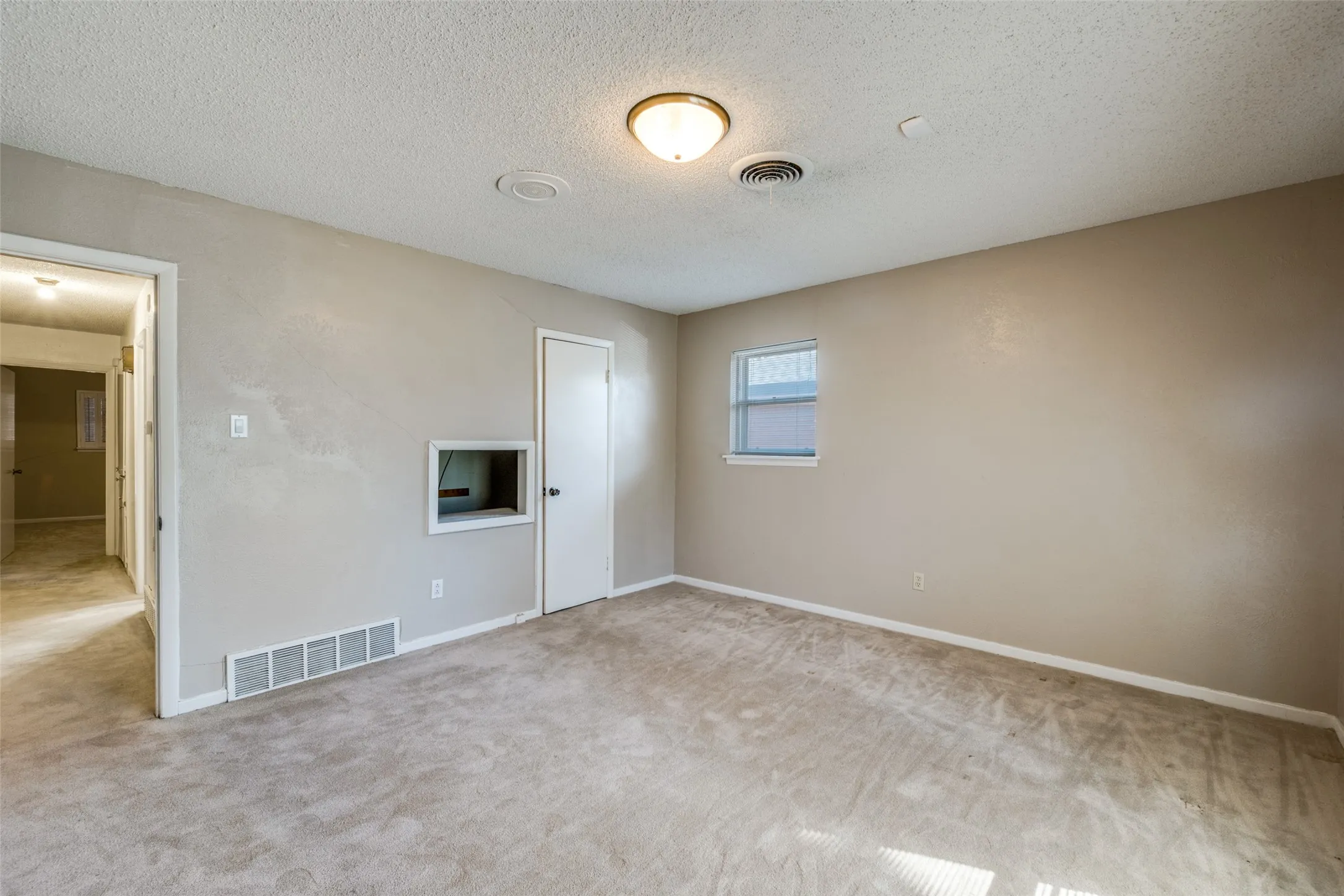 Carpeted room featuring a ceiling-mounted light fixture, a window with white trim, a solid door with a dark knob, and a wall cutout