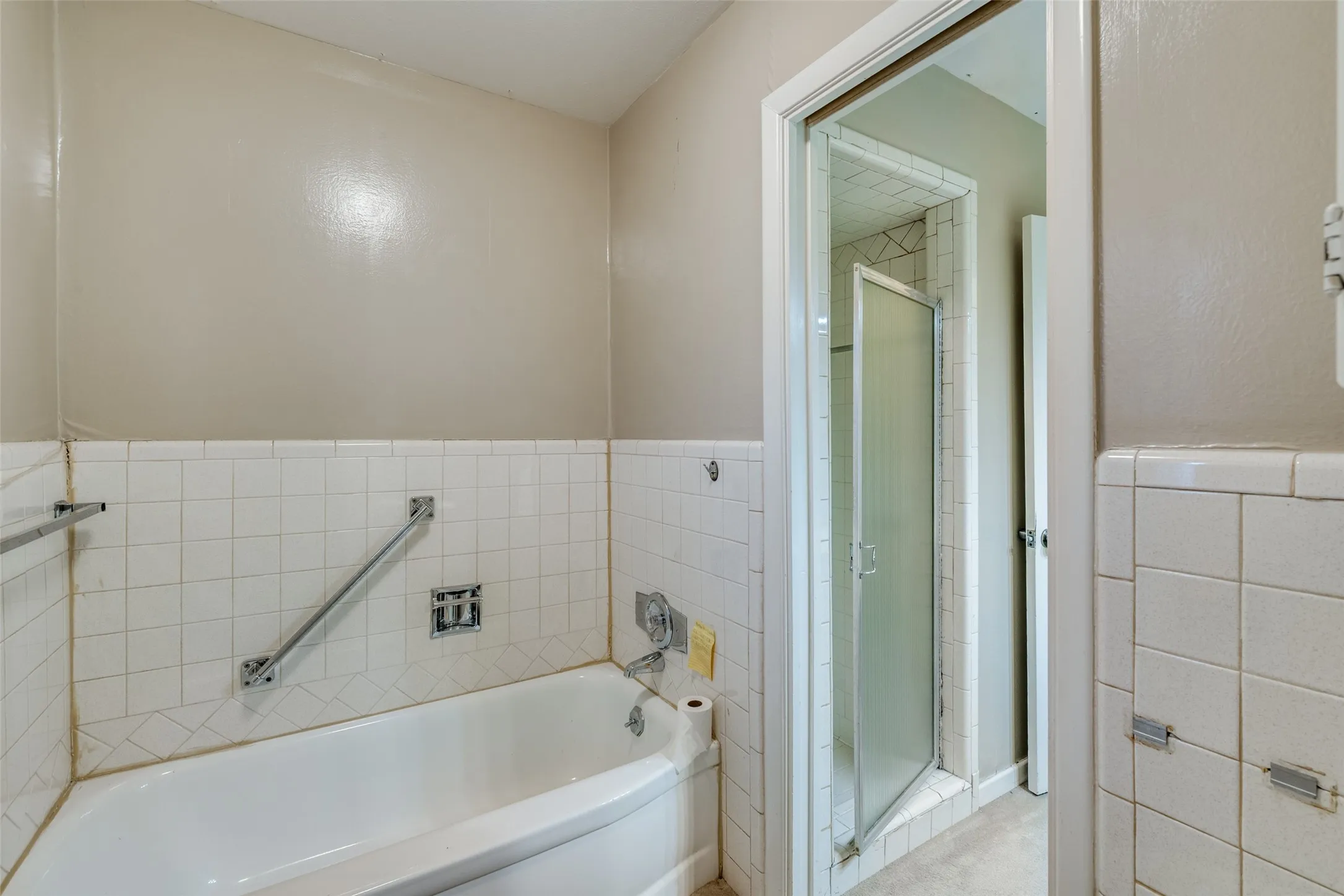 Bathroom featuring a built-in bathtub with white tile surround, chrome grab bars, and an adjacent enclosed shower with a frosted glass door