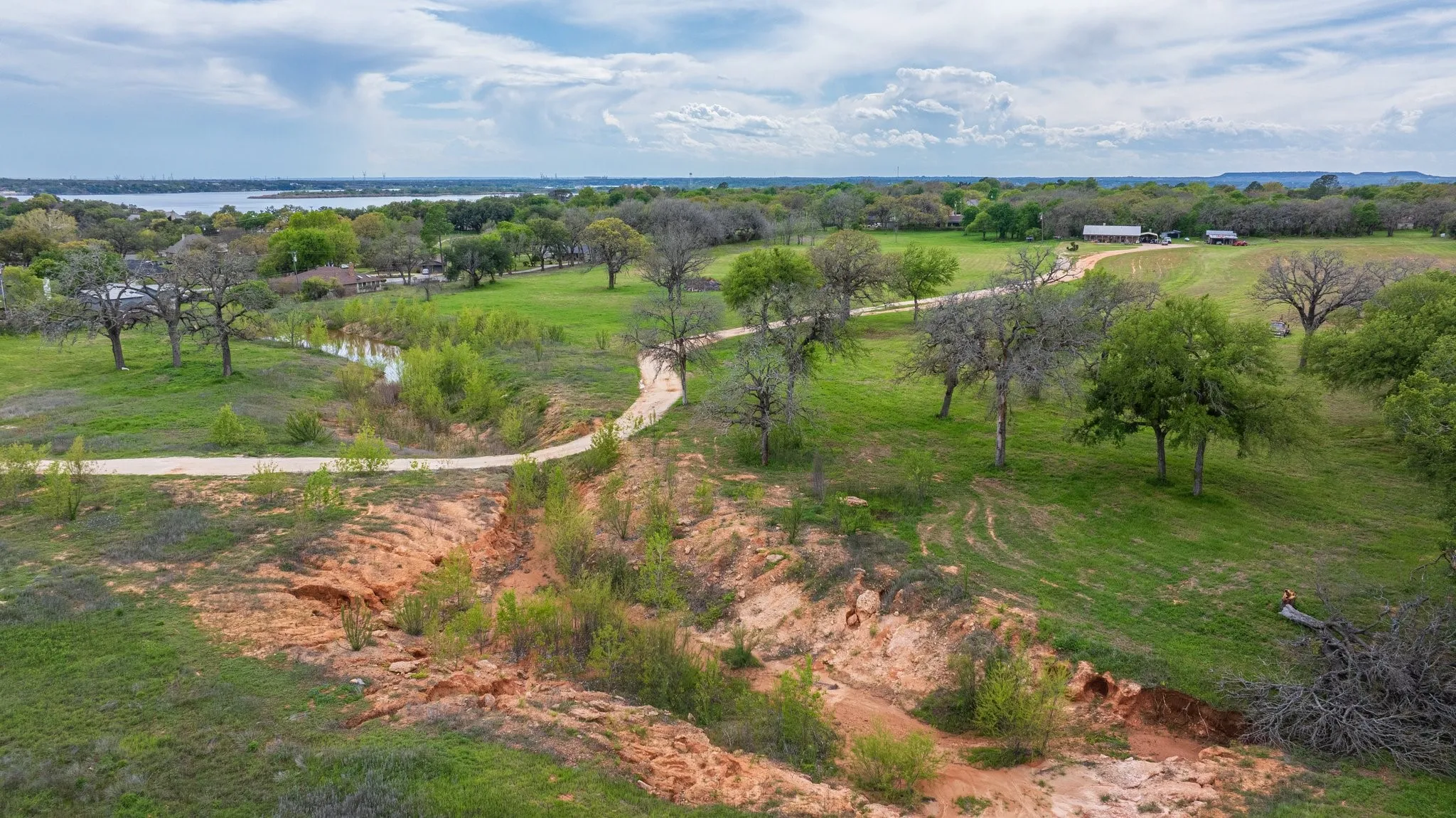 Expansive green acreage featuring mature trees, a winding dirt path, and a distant body of water
