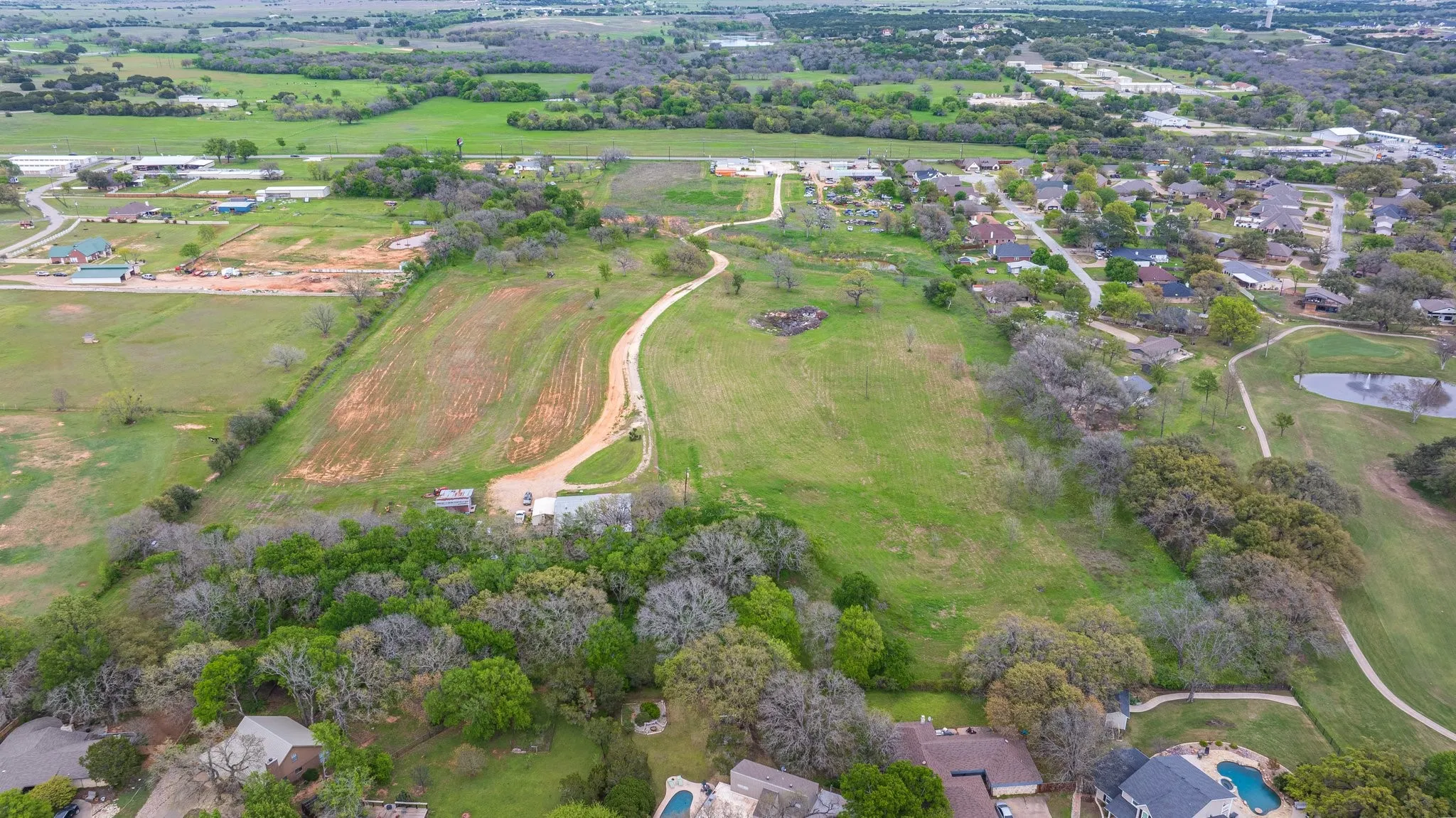 Expansive land parcel featuring a winding unpaved road, mature tree lines, and distant residential areas