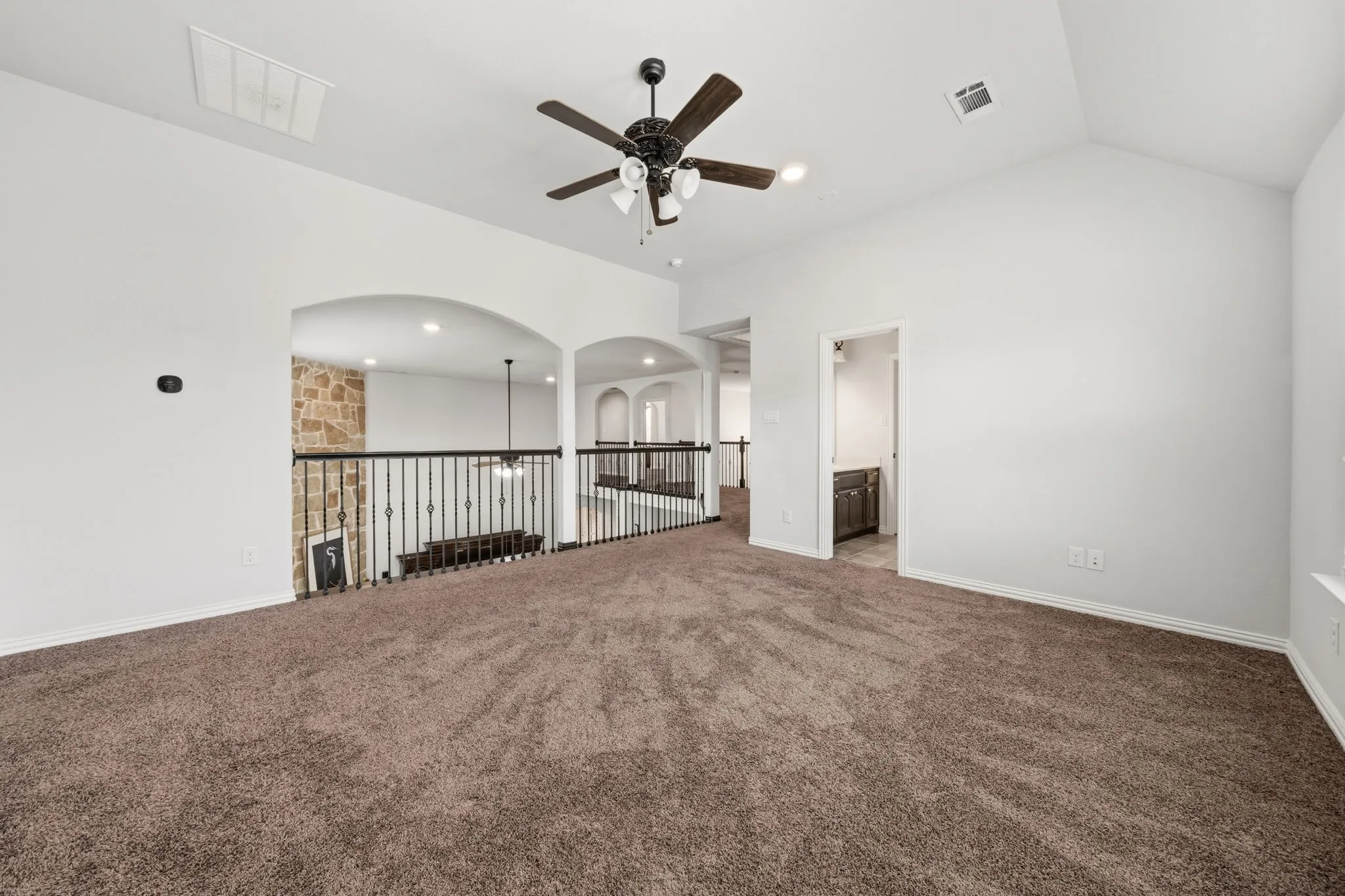 Carpeted open loft area featuring a vaulted ceiling with a ceiling fan, wrought iron railings, stone accent wall, and arched doorways