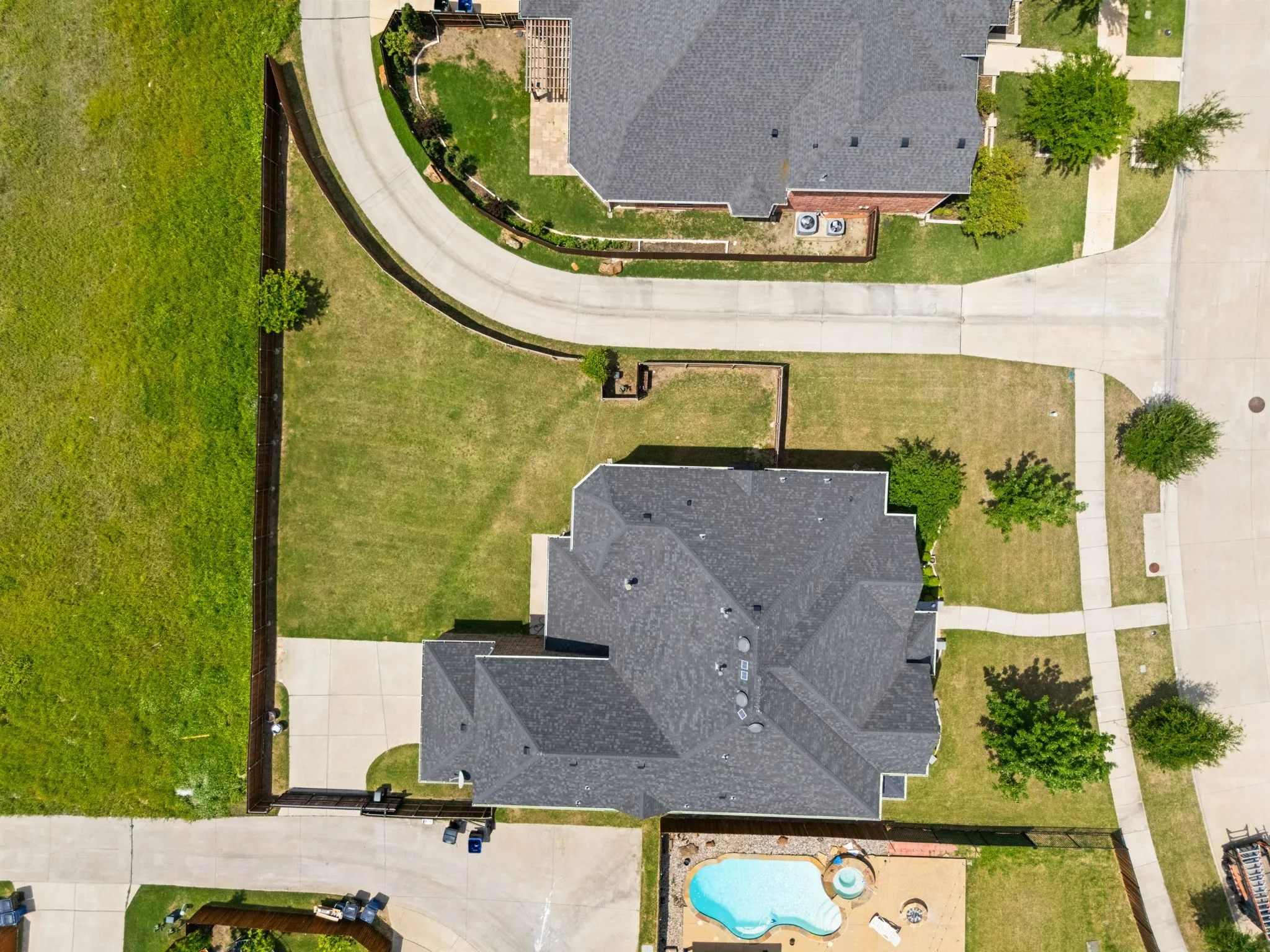 Aerial perspective showcasing a property with a dark shingle roof, extensive manicured lawn, concrete driveway, and a backyard swimming pool with an attached spa