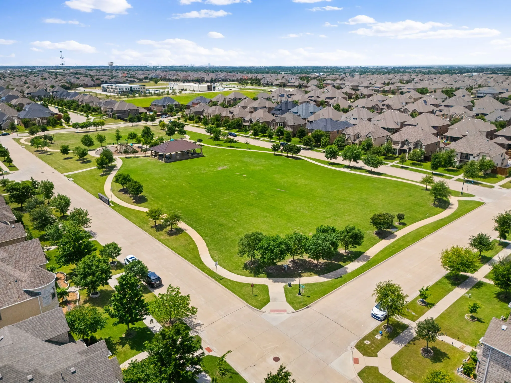 Expansive green space featuring a large manicured lawn, paved walking paths, a covered pavilion, and a playground area with multiple play structures