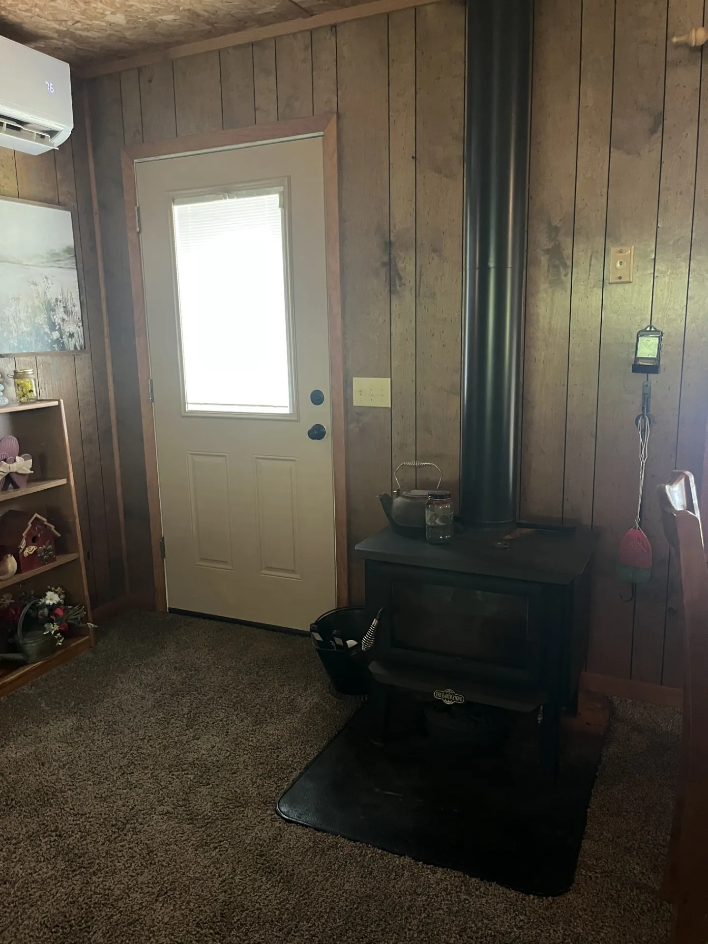 Wood-paneled interior featuring a black wood-burning stove with a protective hearth pad, a white exterior door with an inset window, and a wall-mounted mini-split HVAC unit