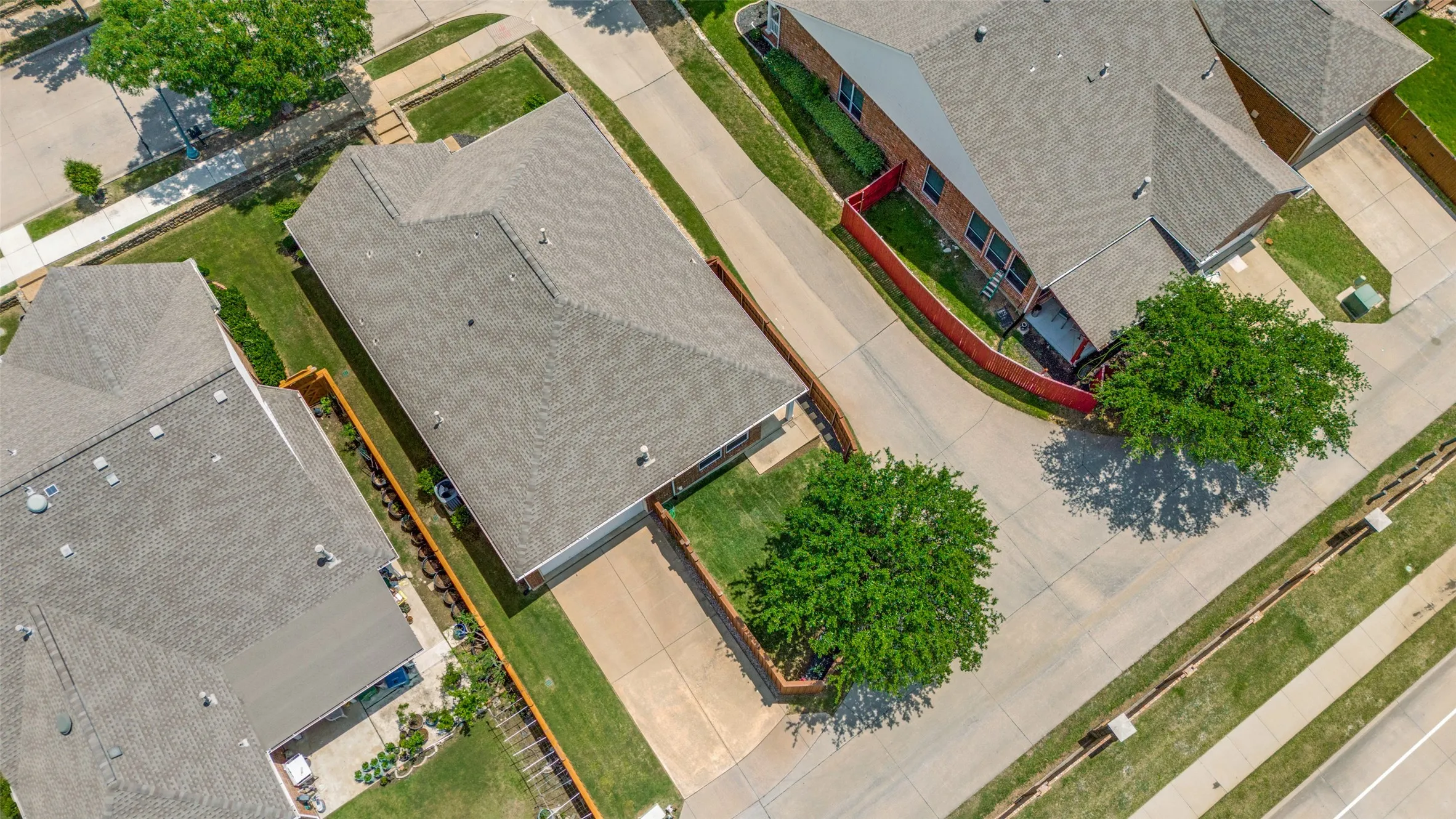 Aerial view of the property and surroundings, featuring a gabled roof, paved driveway, mature tree, and fenced backyard.