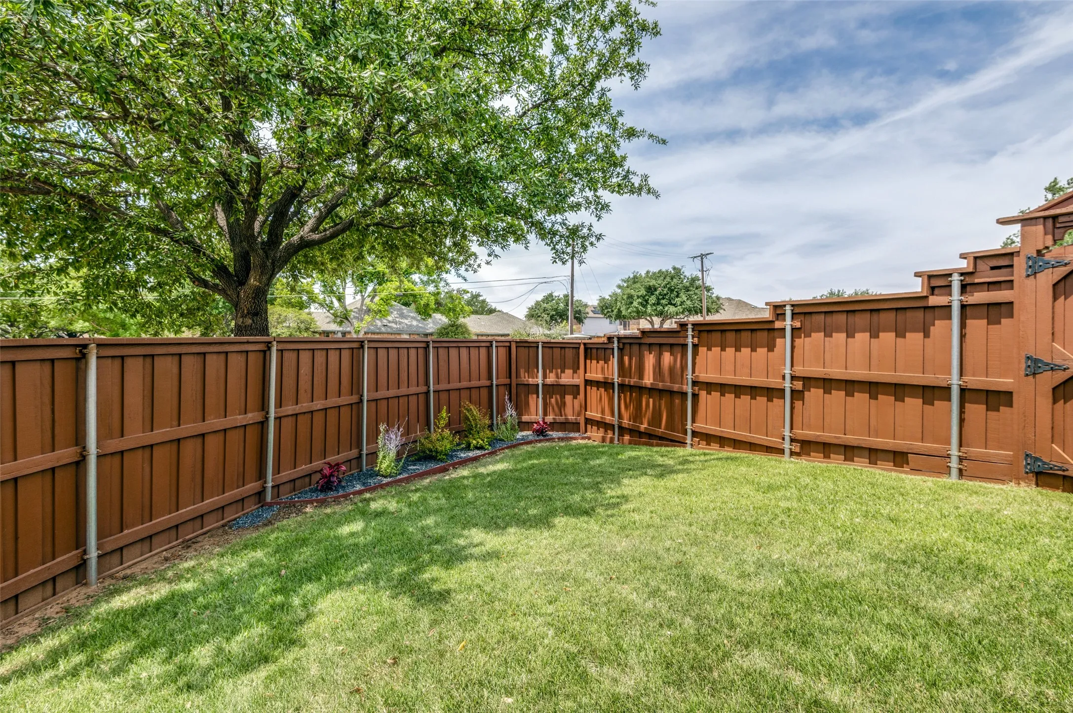 Grassed yard area featuring a stained privacy fence with metal posts and newly landscaped flower bed. Great shade tree!