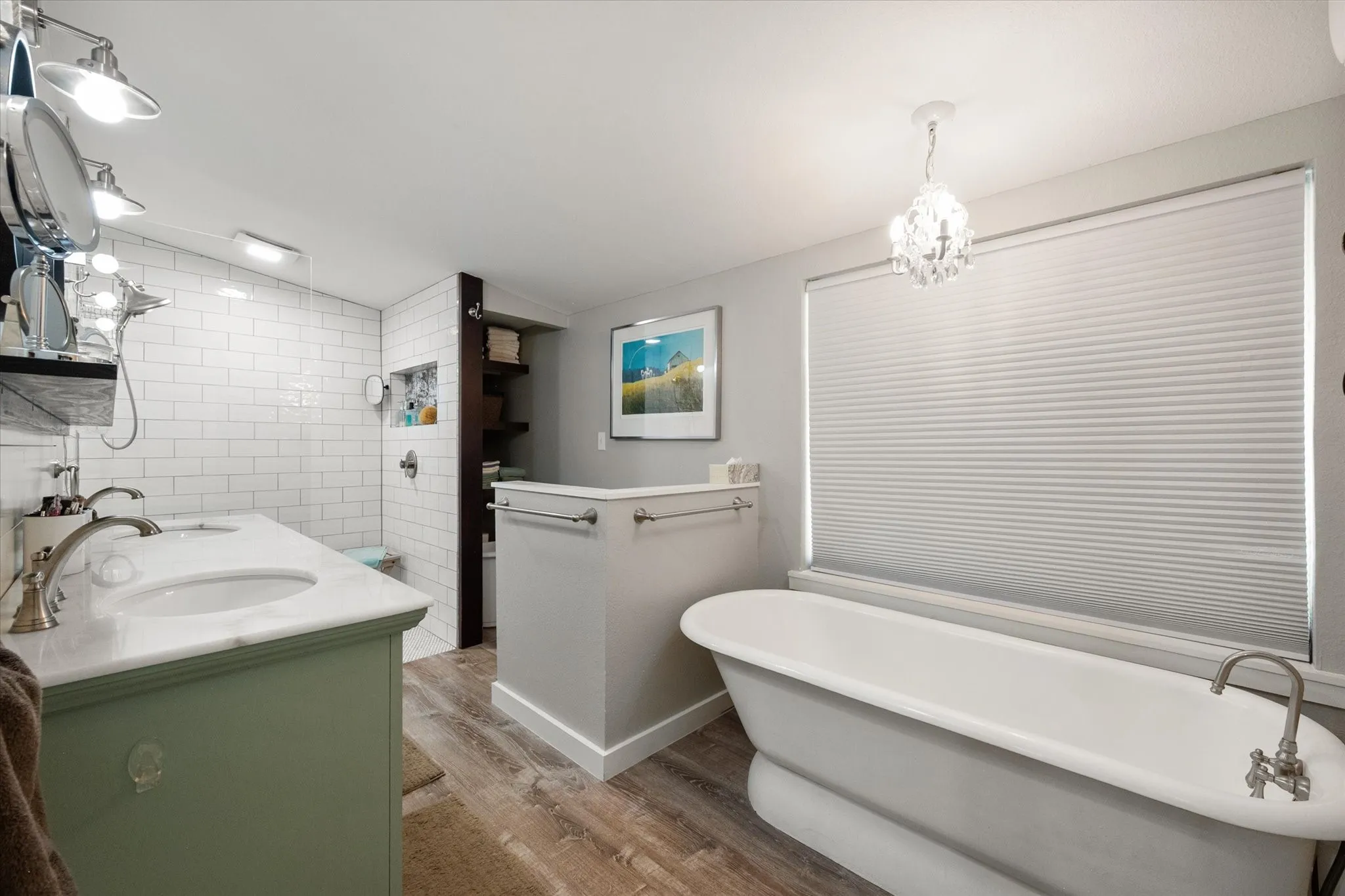 Freestanding bathtub with floor-mounted faucet, wood-finish flooring, dual vanity with undermount sinks, and subway tile shower