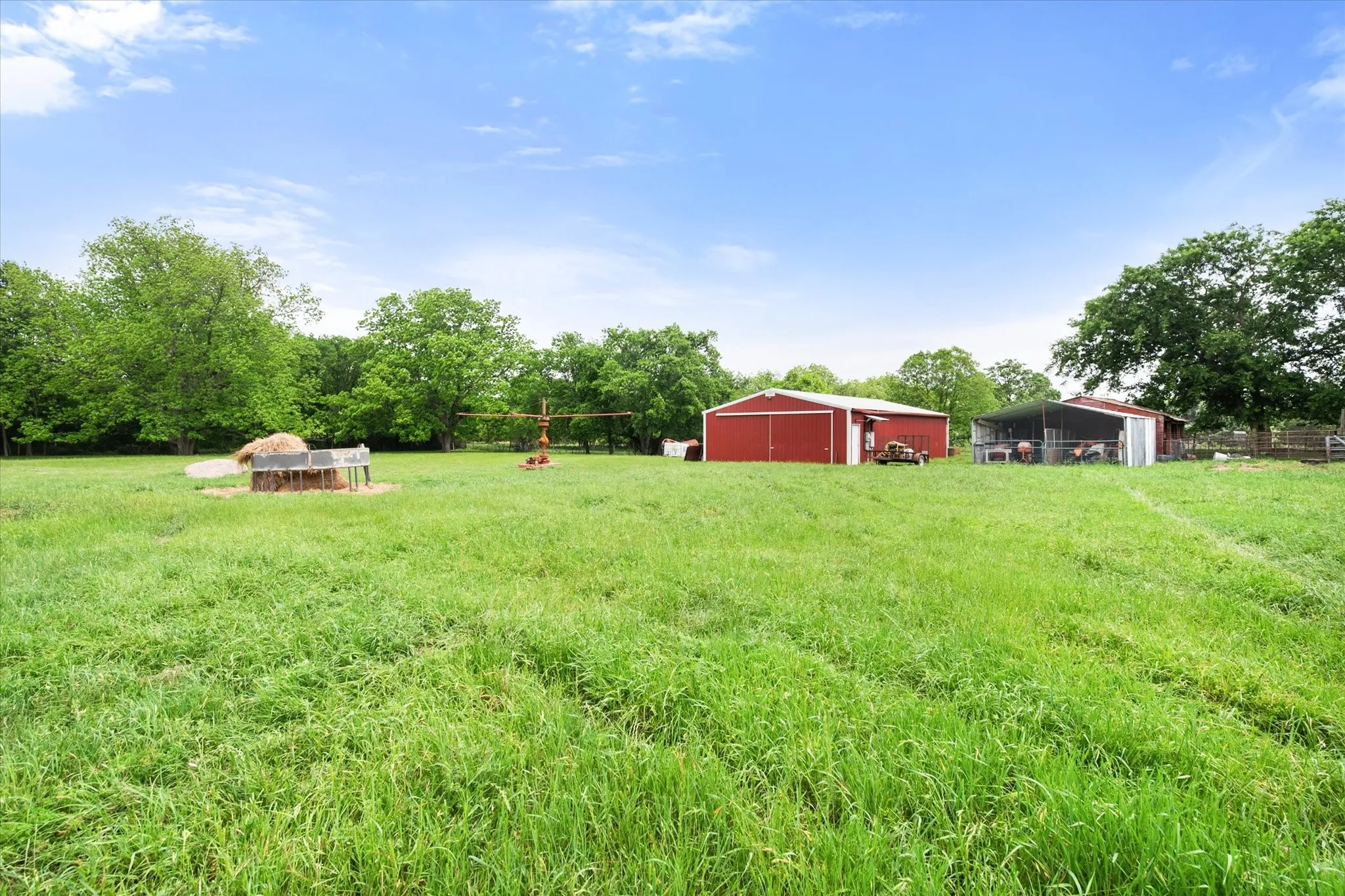 Expansive green pasture with two red agricultural buildings, one enclosed and one open-sided, surrounded by mature trees