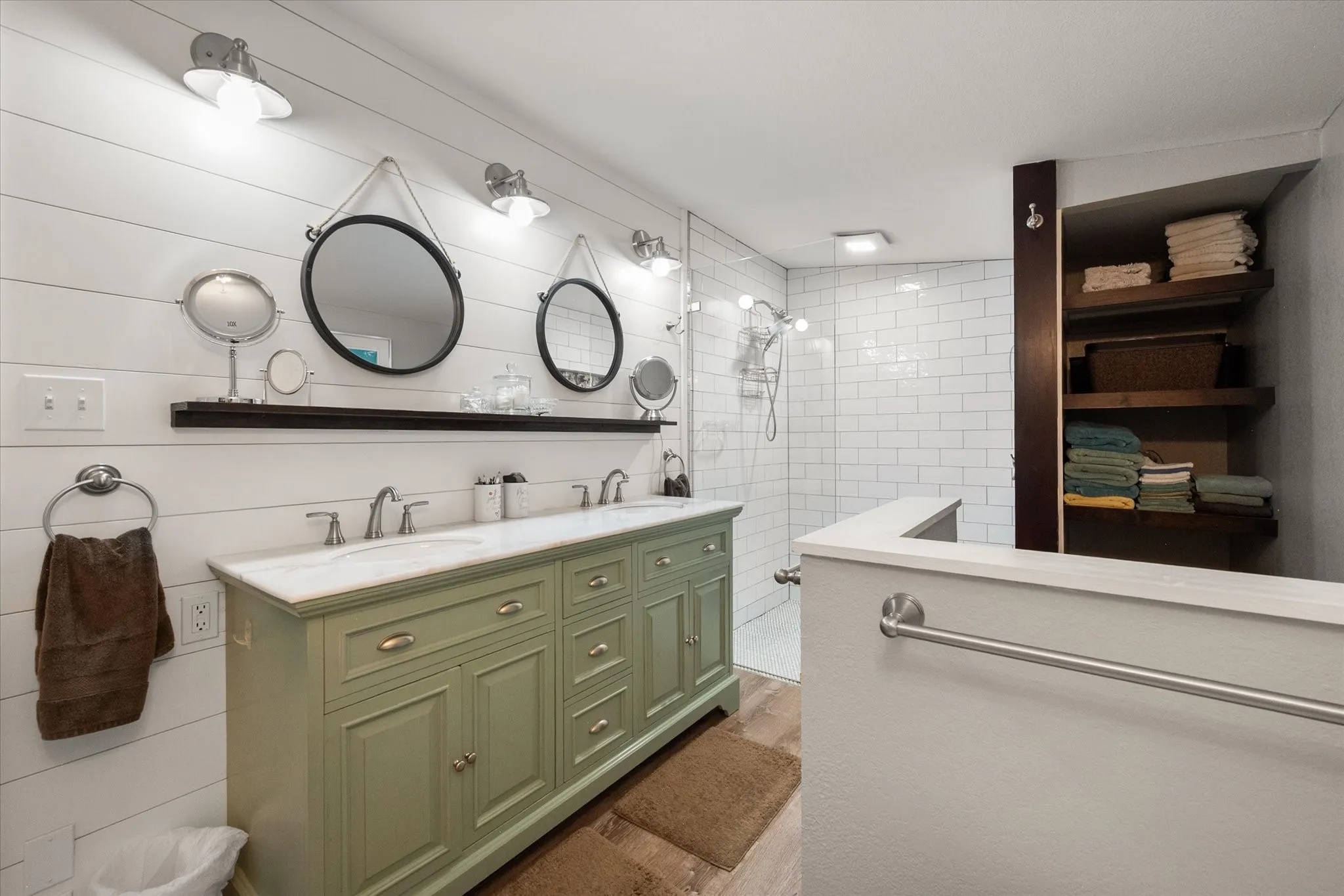 Spacious bathroom featuring a double vanity with a light green cabinet base and dual undermount sinks, white shiplap wall paneling, a glass-enclosed shower with white subway tile, and built-in wood shelving for storage