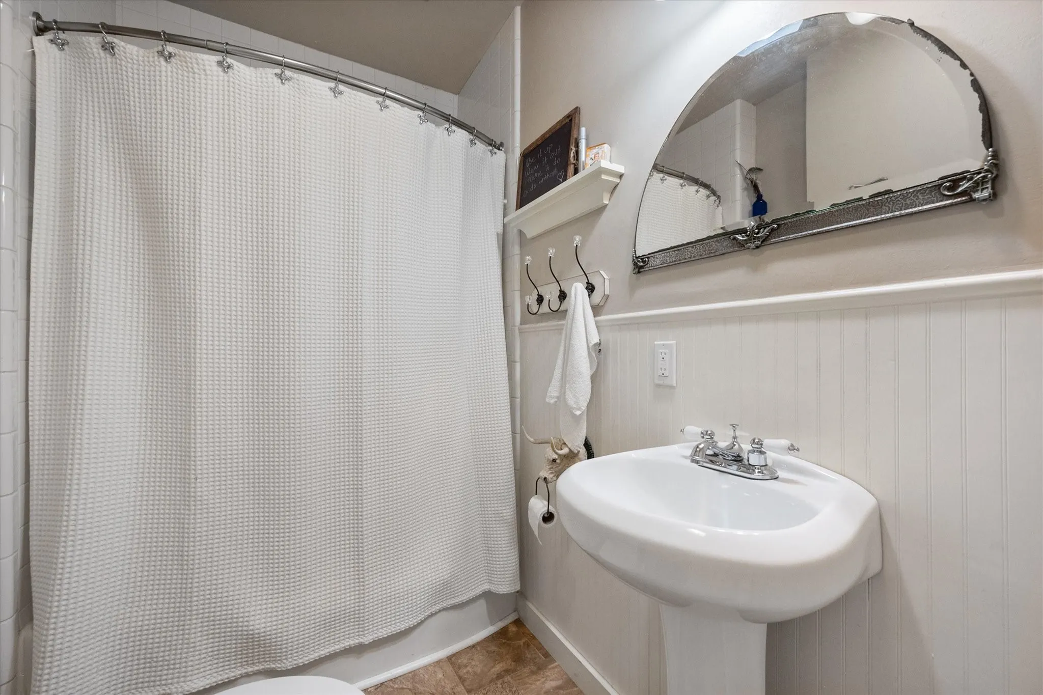 Bathroom featuring a pedestal sink with chrome faucet, wainscoting, and an arched mirror with decorative frame