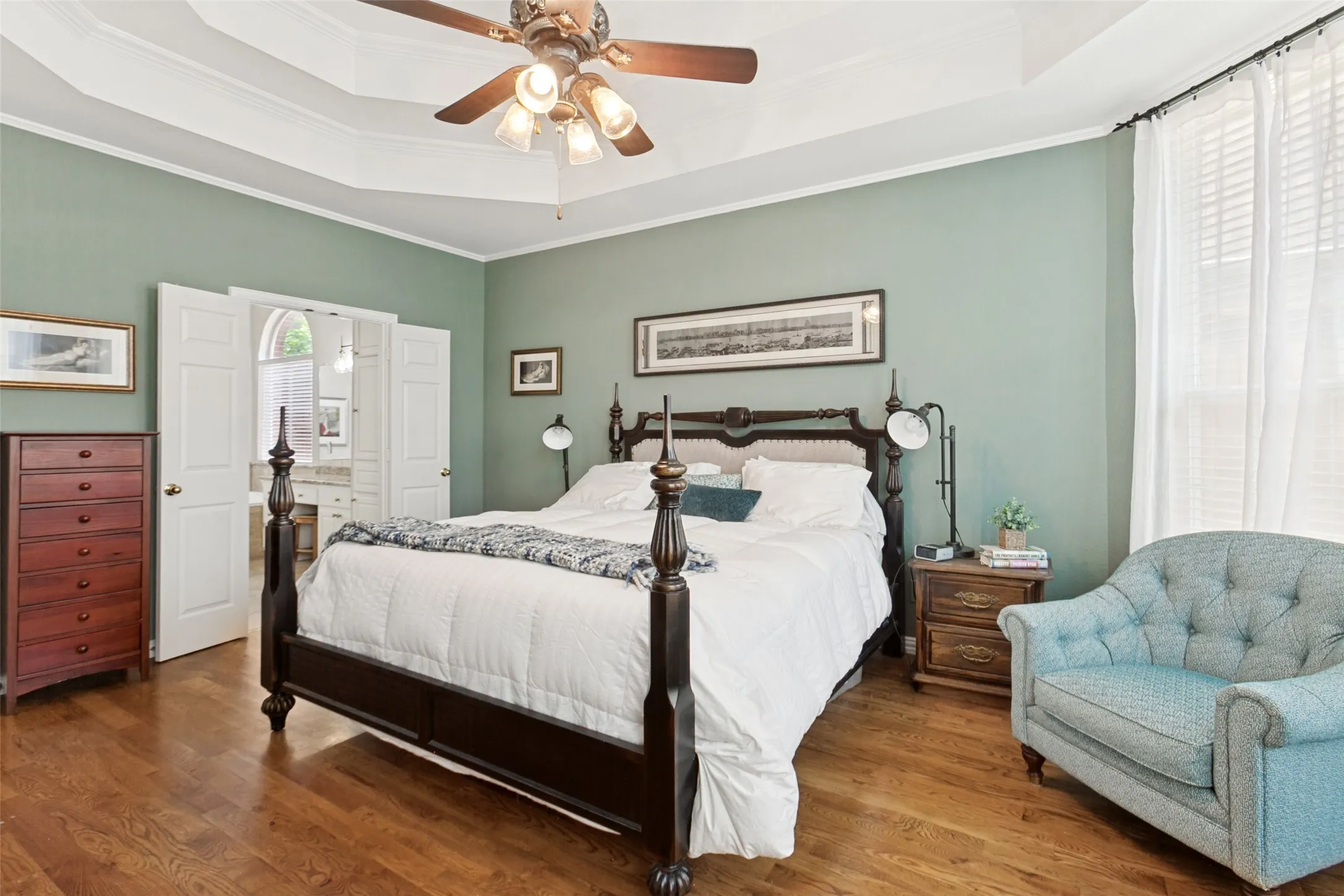 Private first floor primary bedroom suite featuring wood-finish flooring, a tray ceiling with a built-in fan light fixture, and white double doors opening to a spacious bathroom enhanced with a large walk-in closet
