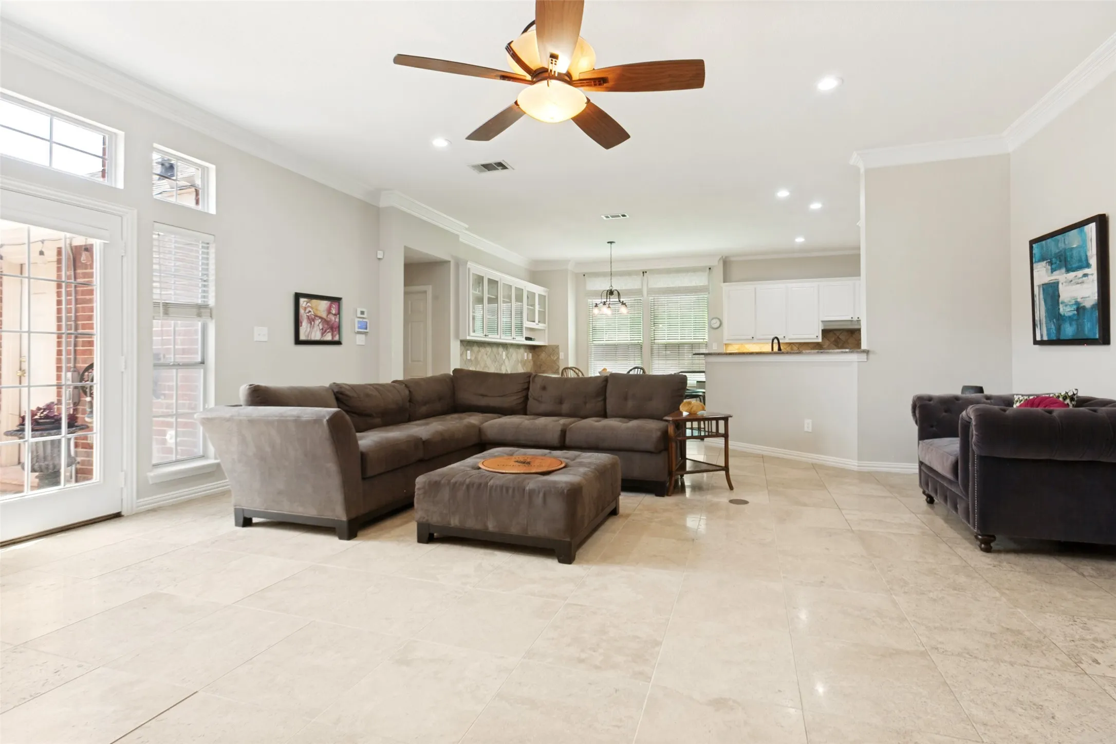 Expansive living area featuring light-toned tile flooring, a ceiling fan with integrated lighting, recessed lighting, crown molding, and tall windows with transom details