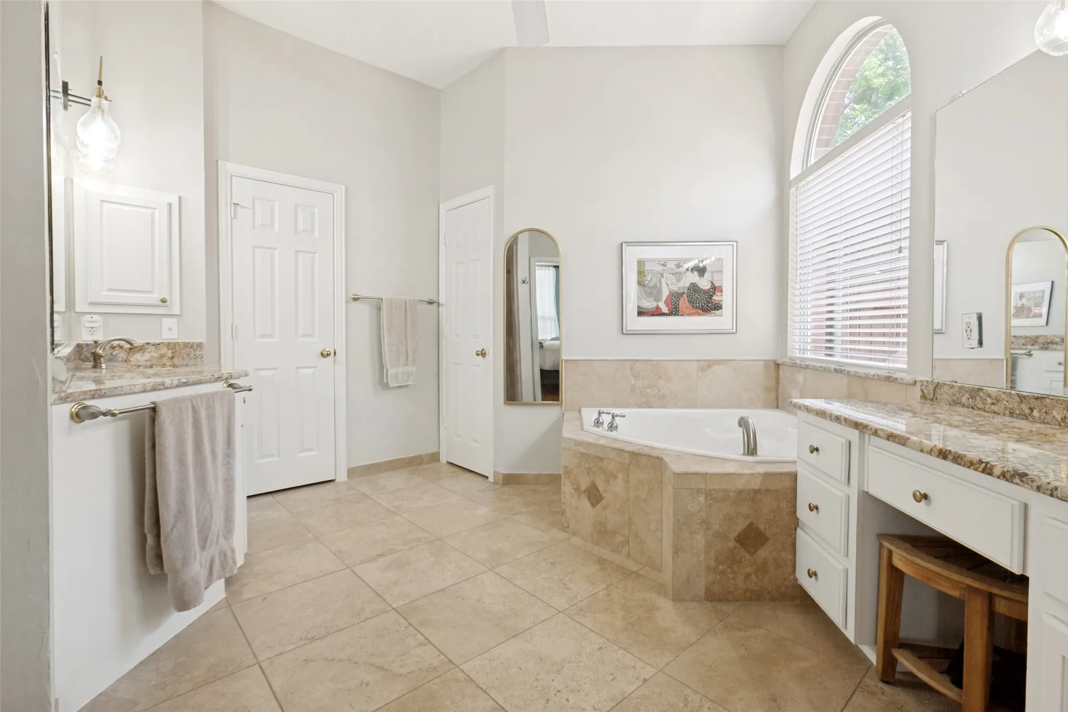 Spacious bathroom featuring a corner jetted tub, arched window, dual vanity areas with granite countertops, walk-in closet, and tile flooring