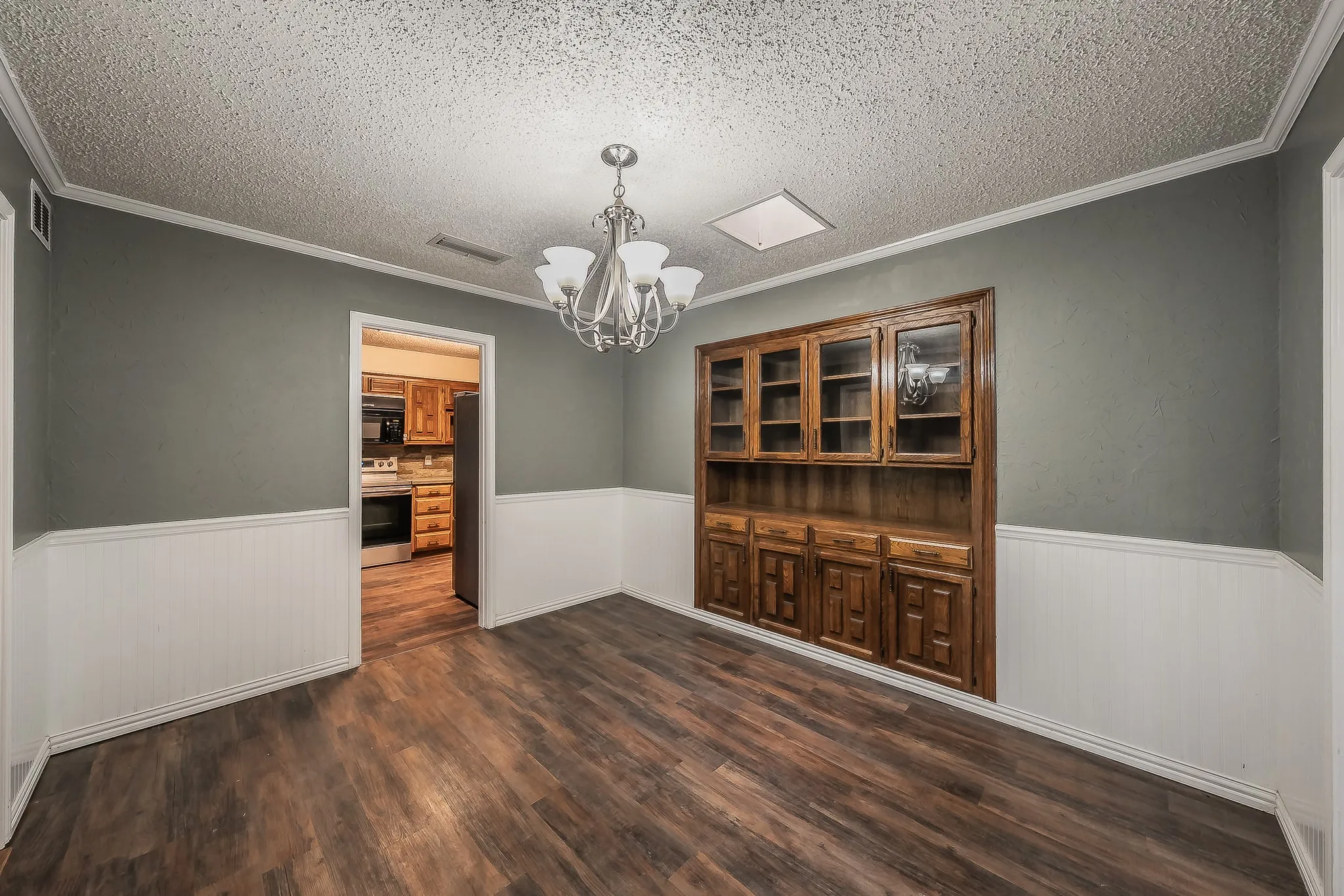 Spacious room featuring wood-finish flooring, white wainscoting, and a built-in wood cabinetry unit with glass-front uppers