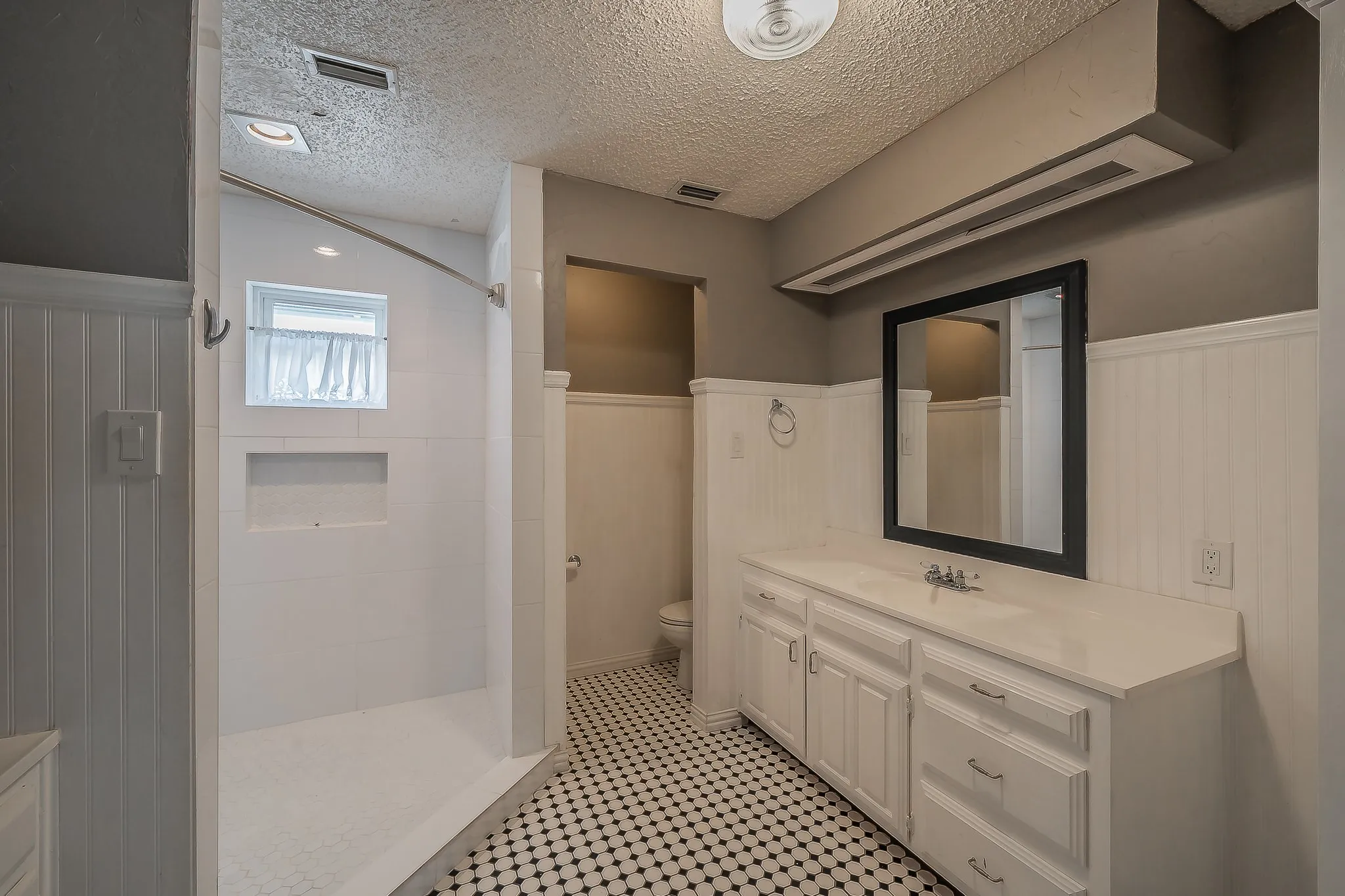 Bathroom featuring a dual-sink vanity with white cabinetry, a black-framed mirror, a walk-in shower with a built-in niche, black and white mosaic tile flooring, and white wainscoting