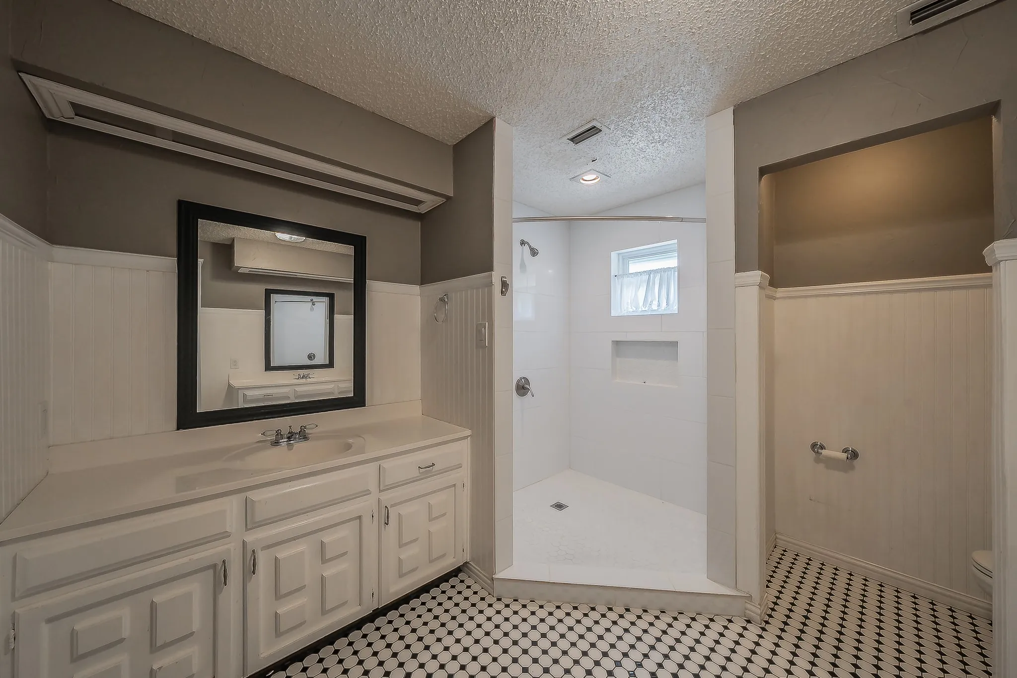 Bathroom featuring a white vanity with integrated sink, black framed mirror, white beadboard wainscoting, and black and white hexagonal floor tiling