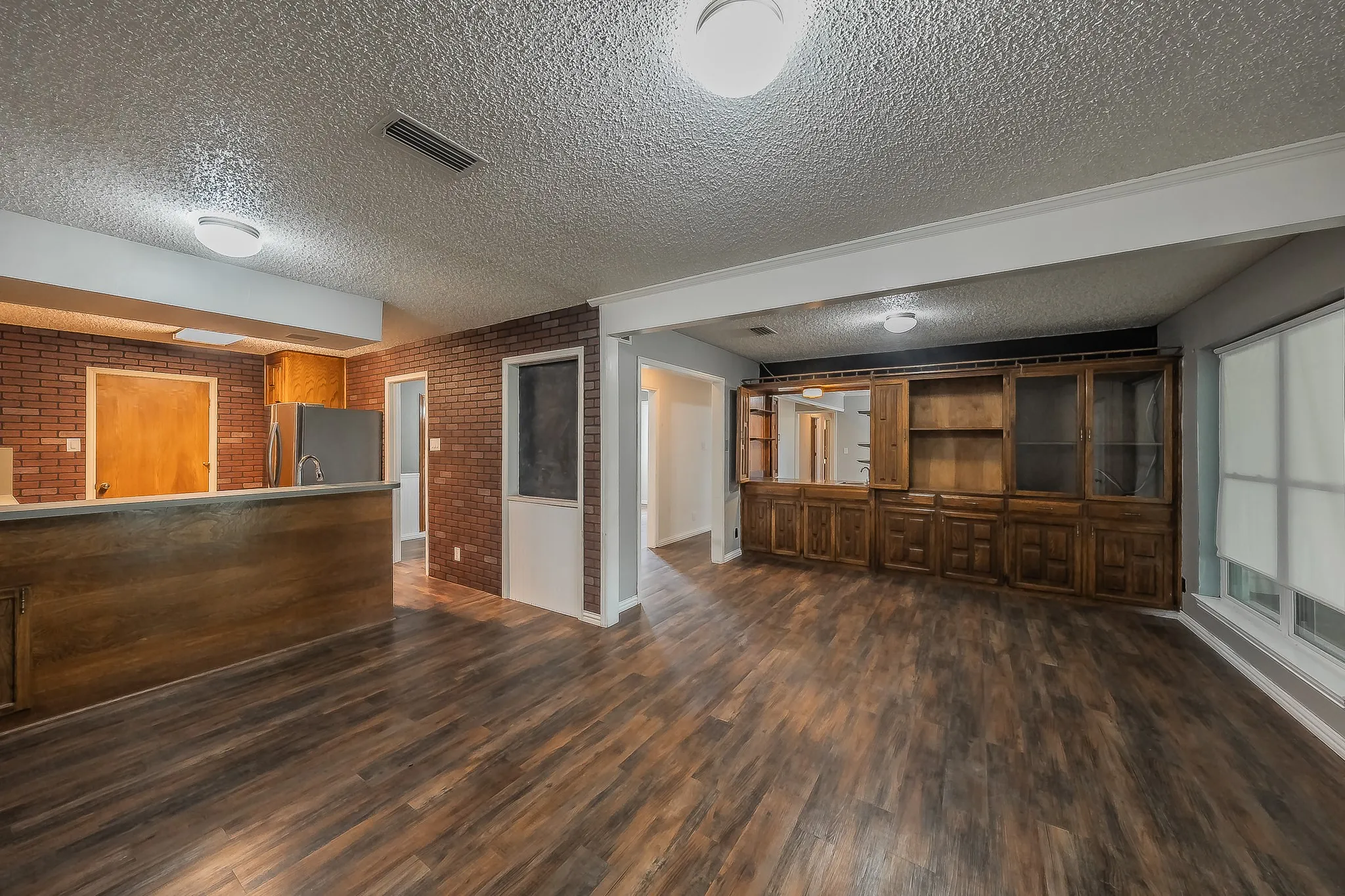 Expansive main living area featuring wood-finish flooring, a built-in wooden entertainment center with glass-front cabinetry, a window with roller shades, and a partial brick wall accent