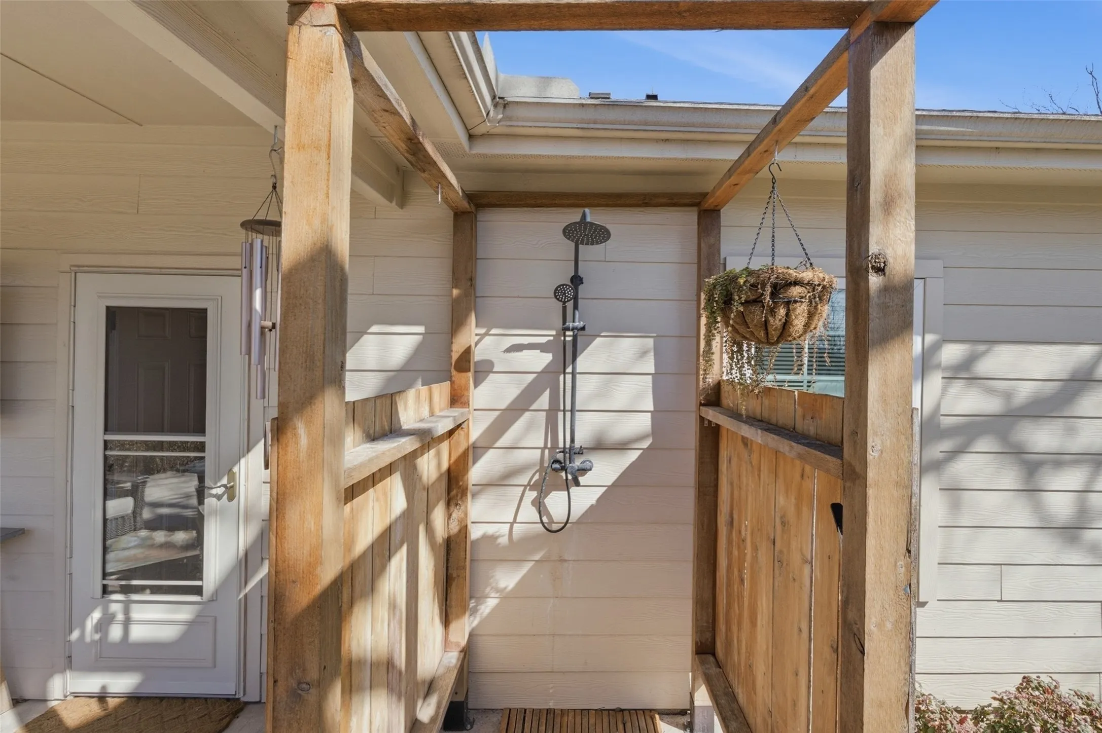 Outdoor shower enclosure with natural wood framing and slatted privacy walls, featuring a multi-function shower system and white horizontal siding backdrop