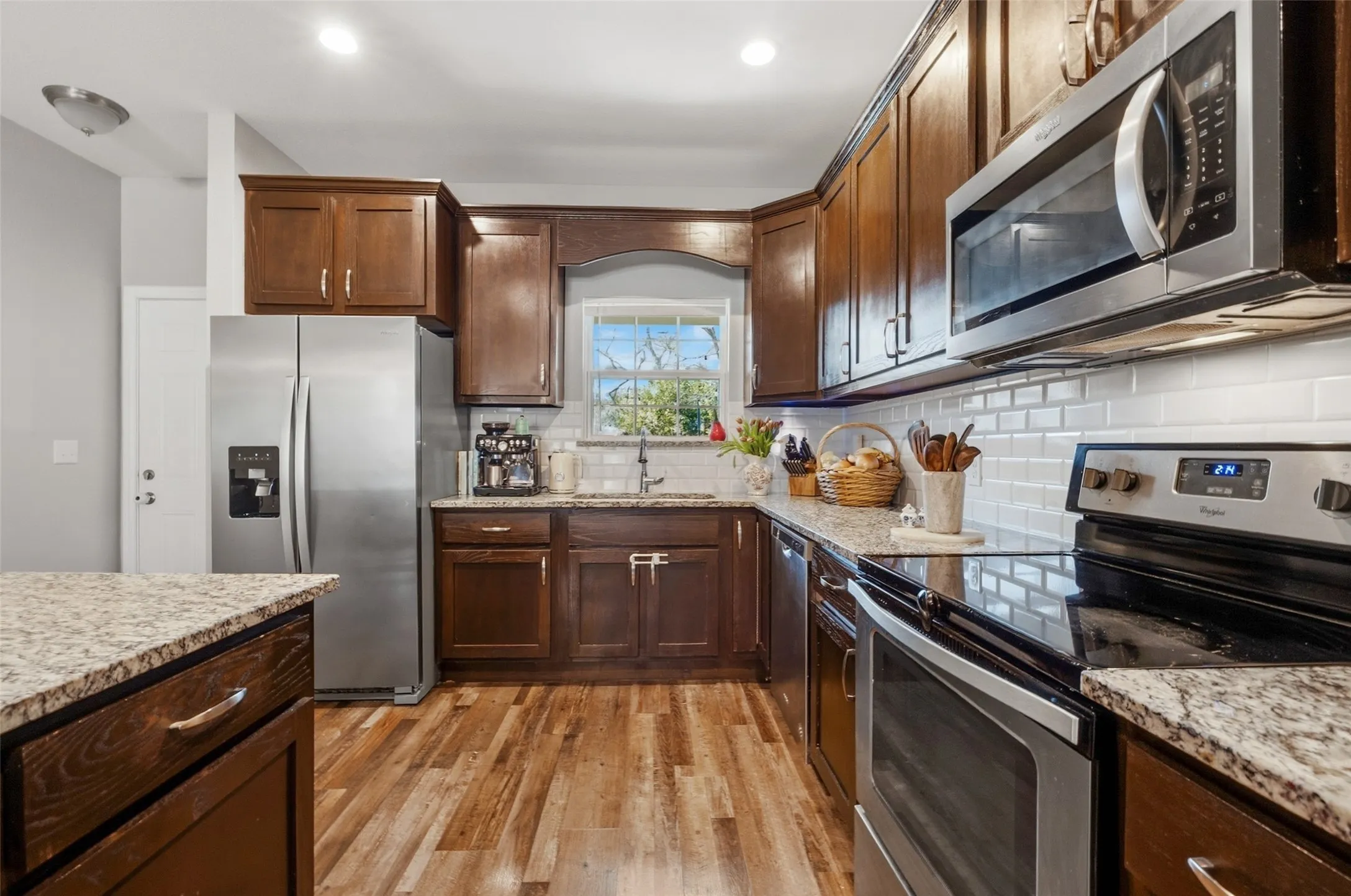 Kitchen featuring dark wood cabinetry, stainless steel appliances, light granite countertops, white subway tile backsplash, and wood-finish flooring
