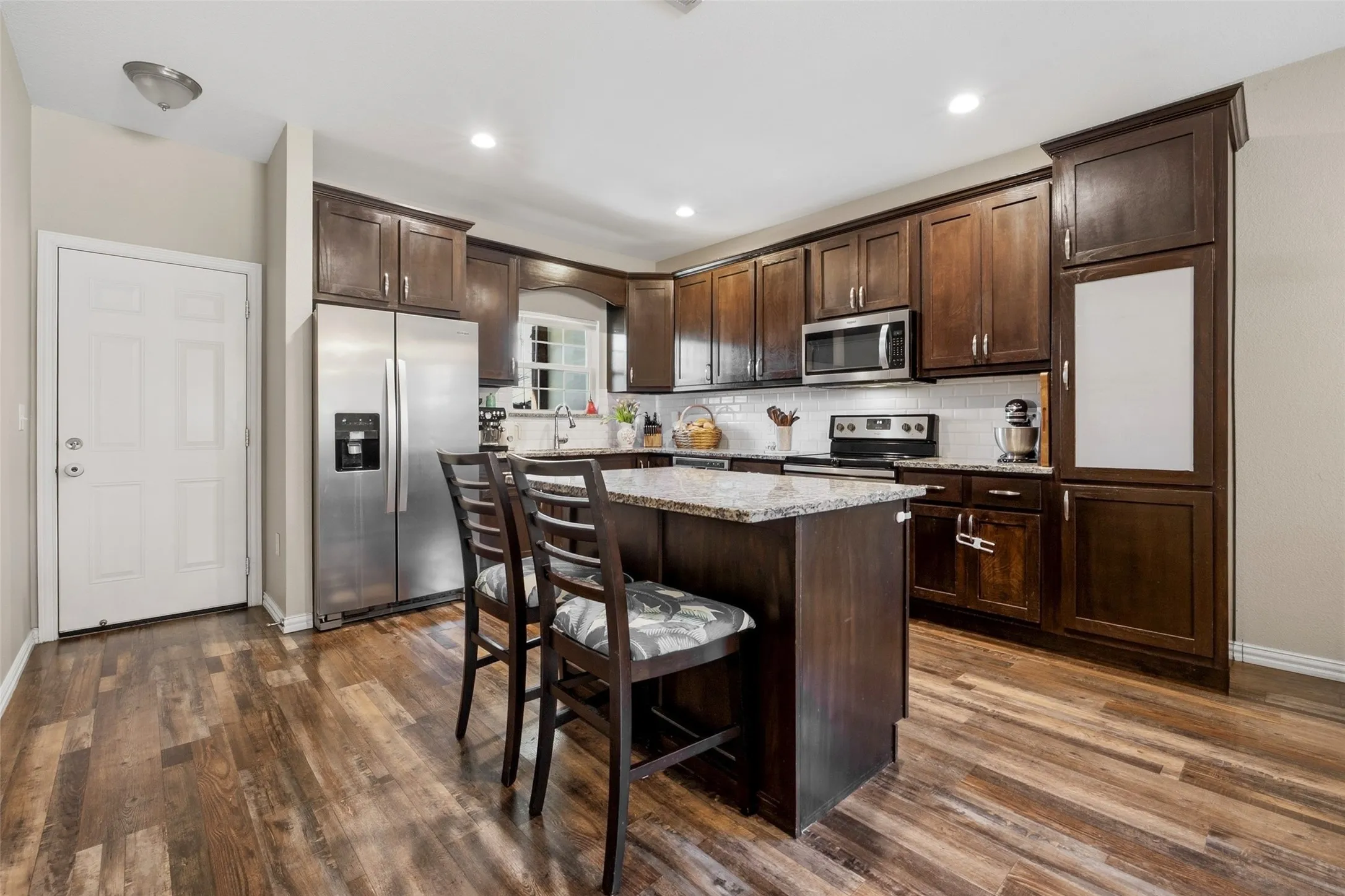 Kitchen featuring wood-finish flooring, dark wood cabinetry, a center island with a granite-style countertop, stainless steel appliances, and a white subway tile backsplash