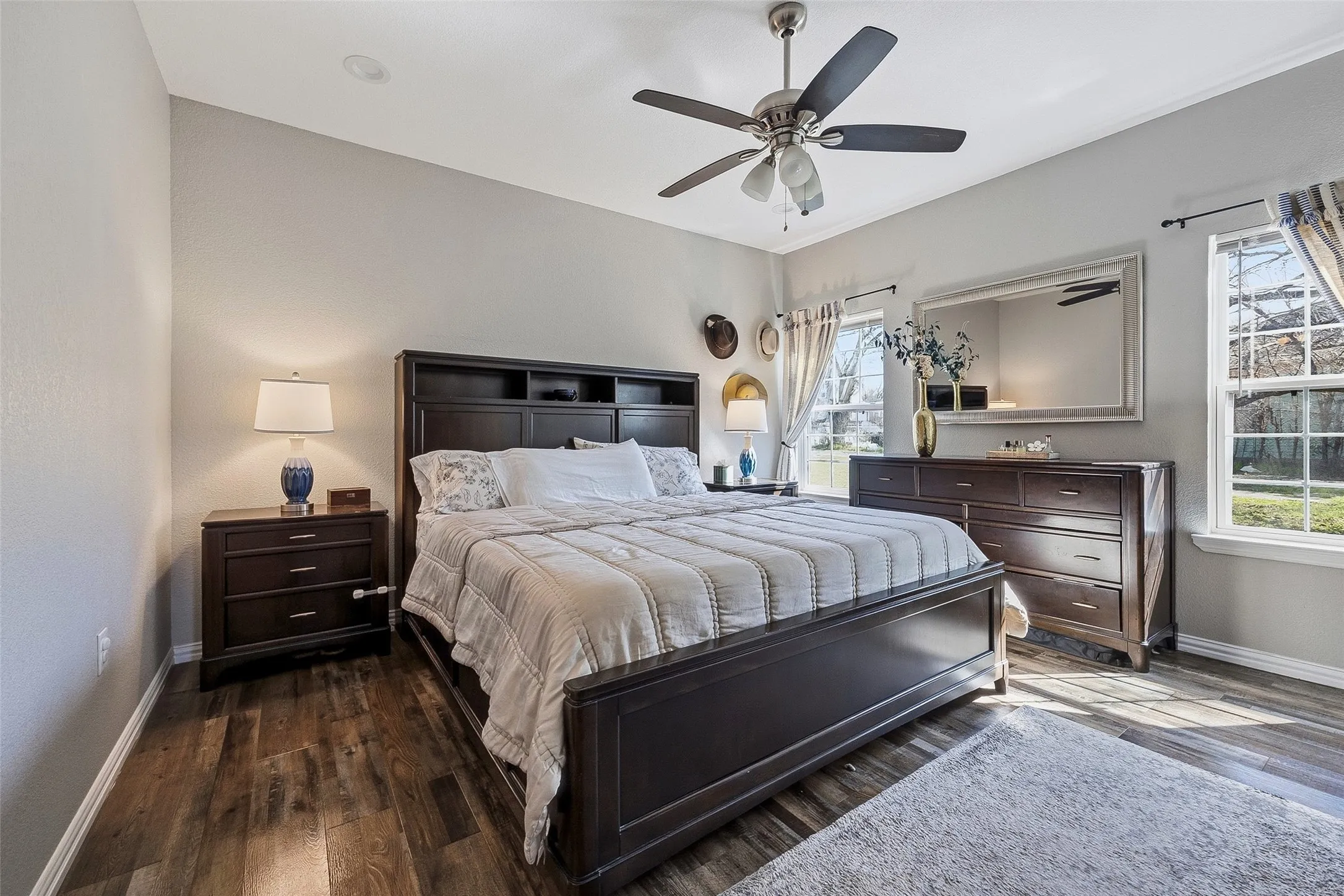 Spacious bedroom featuring wood-finish flooring, two large windows, a ceiling fan with integrated lighting, and neutral wall paint