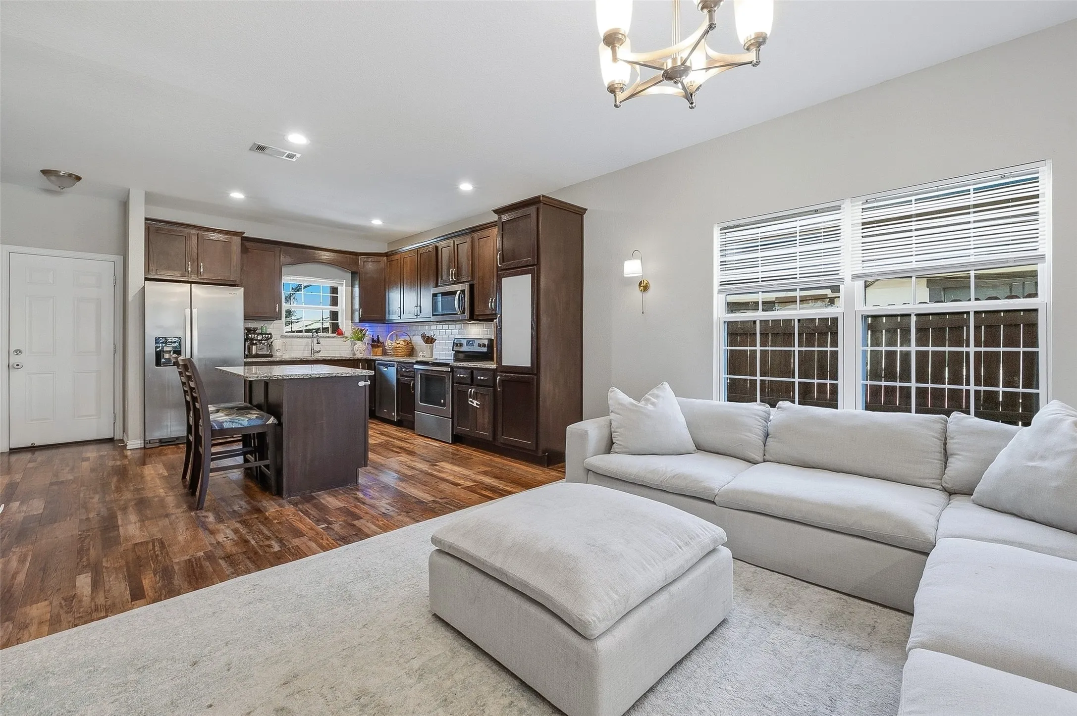 Open-concept living area featuring rich wood-finish flooring, a kitchen island with stone countertop, stainless steel appliances, and a tiled backsplash