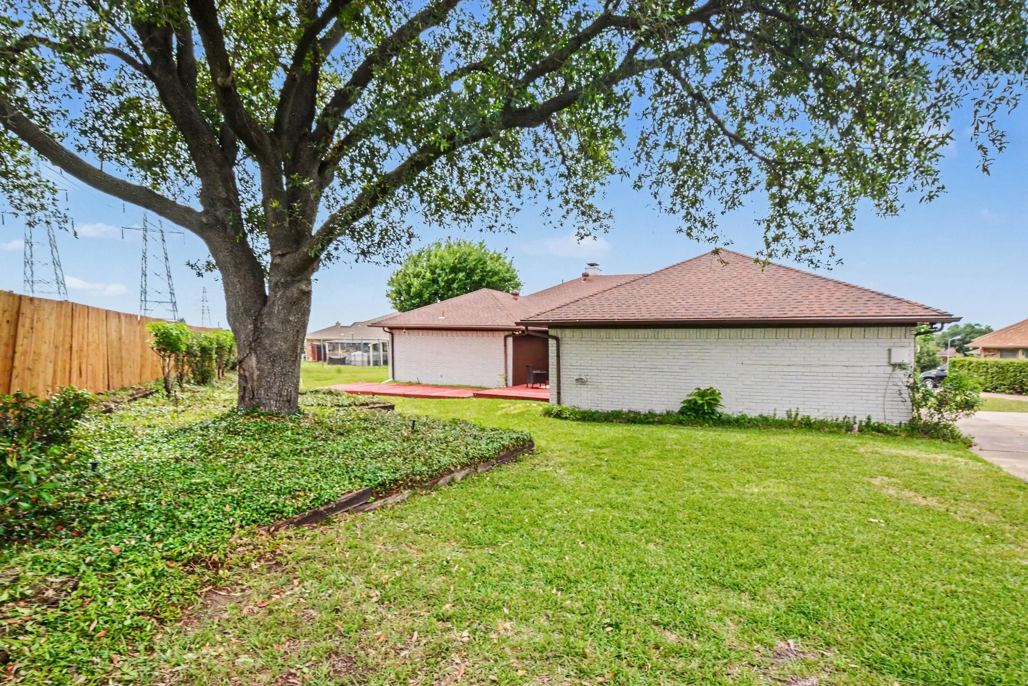Expansive backyard featuring a mature shade tree, manicured lawn, wood fencing, white brick exterior, and a red-painted patio area