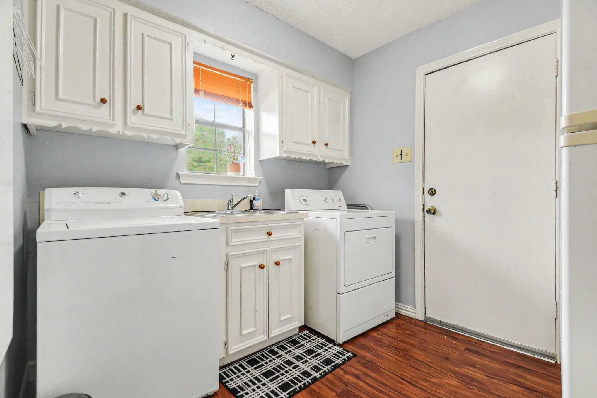 Laundry area featuring wood-finish flooring, white cabinetry with wood knobs, a utility sink, and a window for natural light