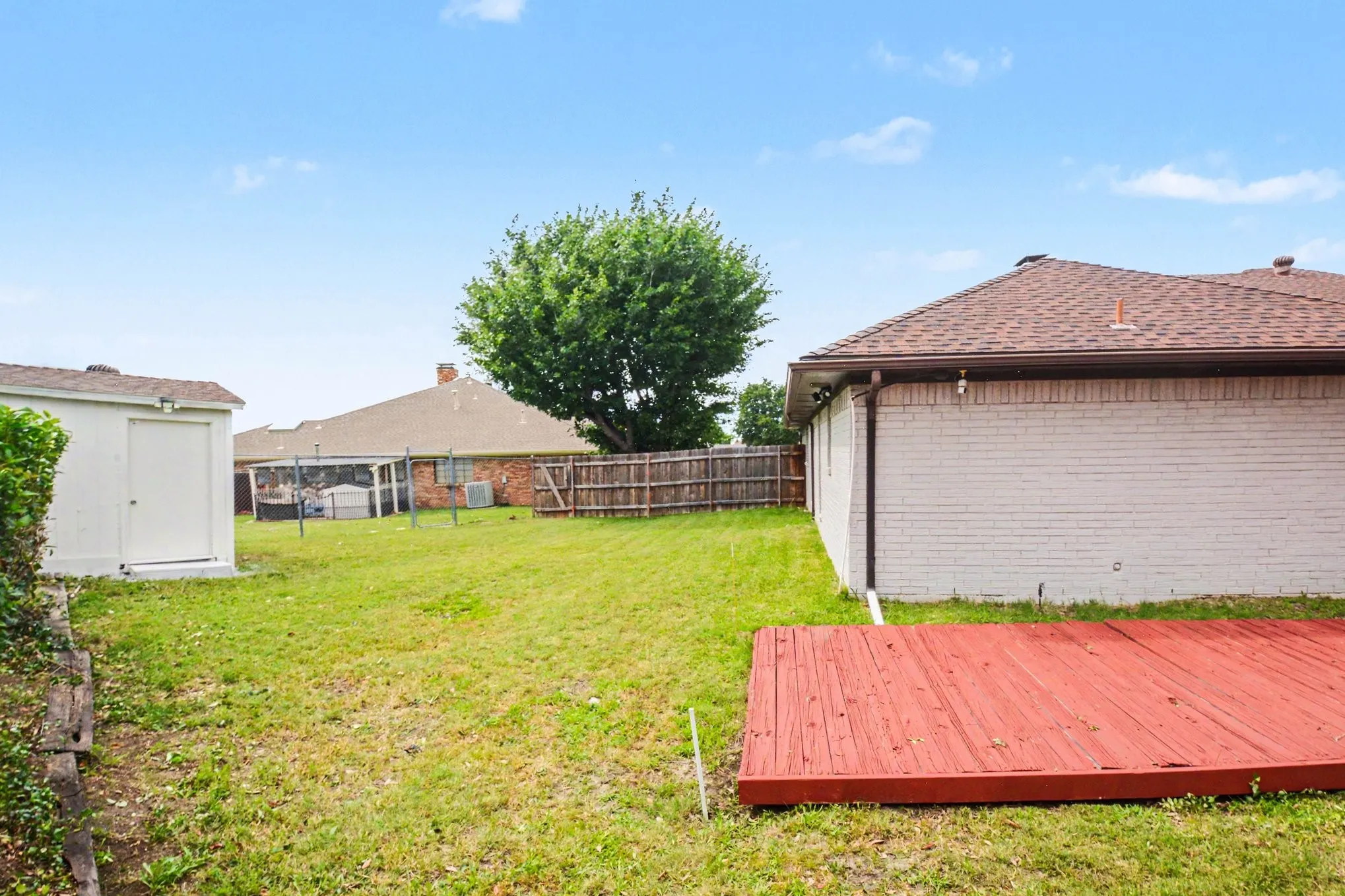 Spacious backyard featuring a red wood-finish deck, a detached white storage shed, and a wood-panel perimeter fence