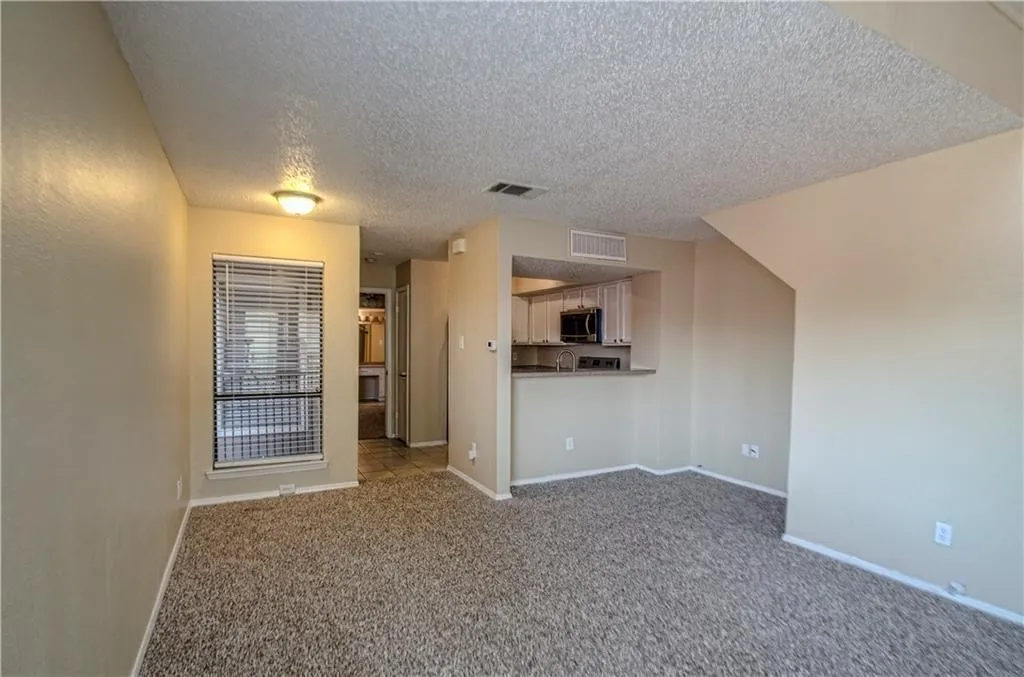 Living area featuring a textured carpet, neutral wall tones, and a window with blinds