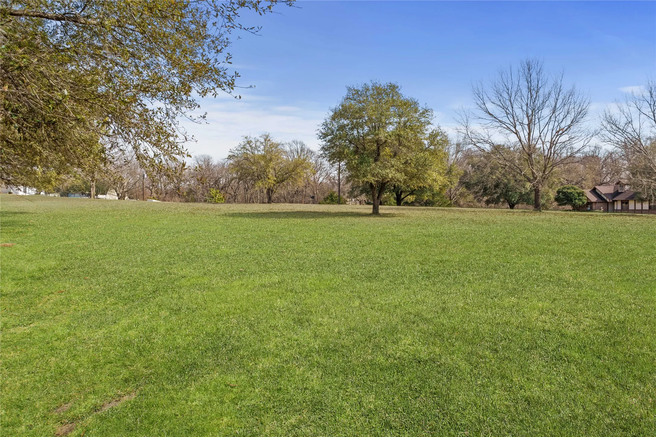 Expansive green lawn with mature trees and a distant building featuring a brown roof and light-colored siding