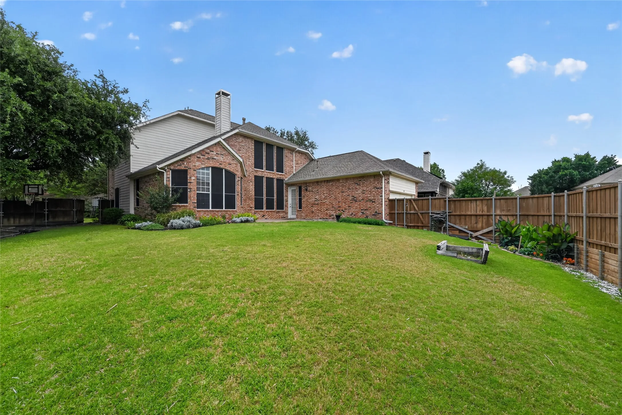 Expansive rear lawn featuring a brick and siding exterior, multiple windows with dark shutters, a prominent chimney, and a wooden privacy fence