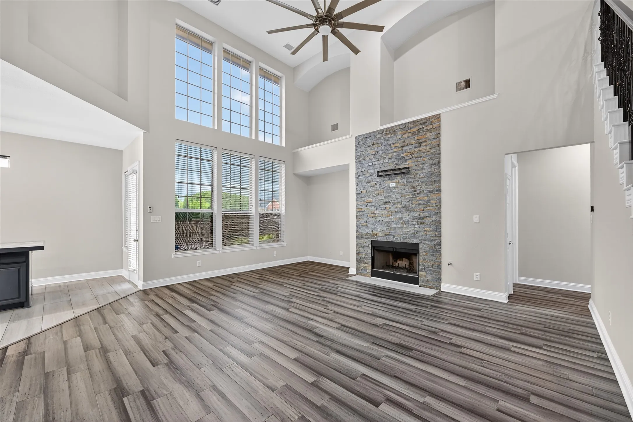 Spacious living area featuring high ceilings, stacked stone fireplace, wood-finish flooring, a contemporary ceiling fan, and multiple large windows